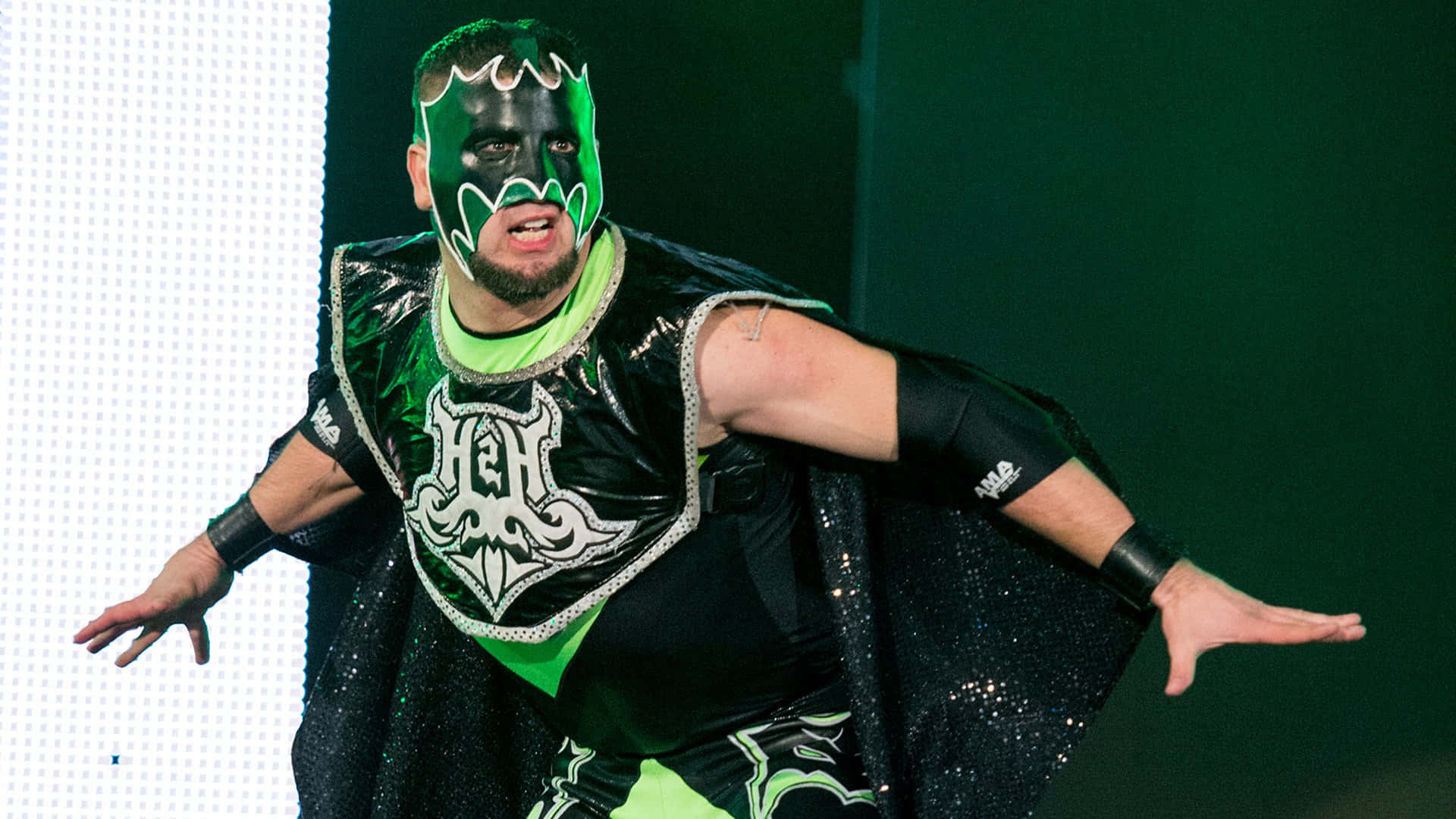 WWE 桌布 壁紙 — 戴著綠色和黑色面具、身穿閃亮服裝的摔角手在明亮的舞台燈光下擺出戲劇性的姿勢。