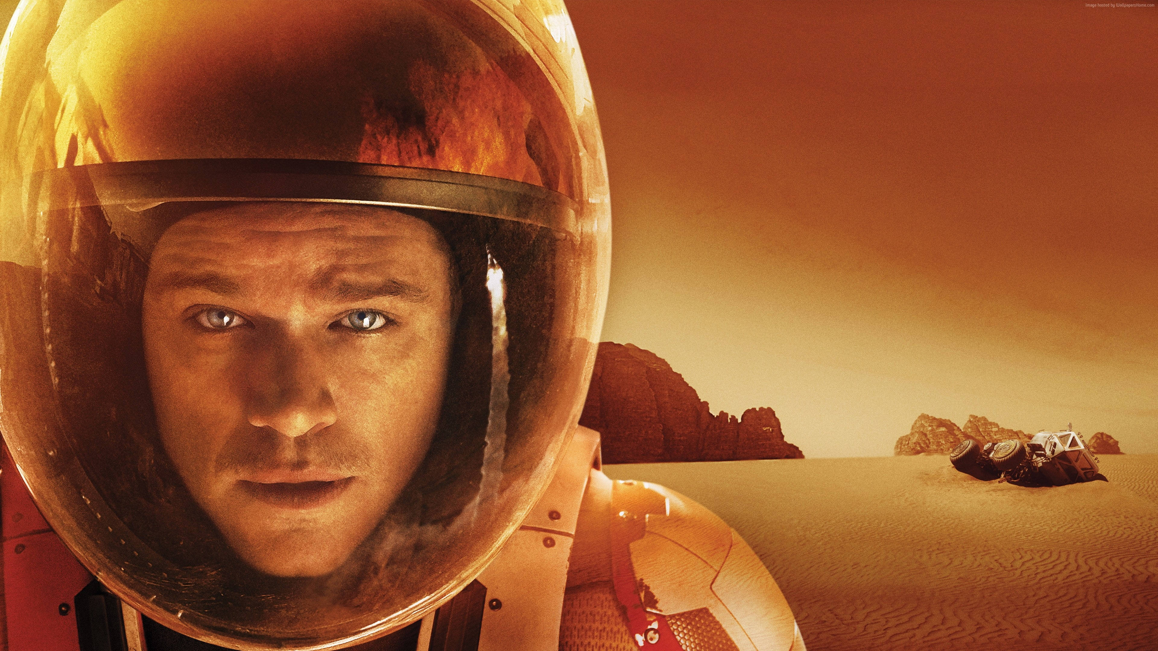 Matt Damon 桌布 壁紙 — 戴著頭盔的太空人從火星表面凝視，背景有探測車和岩石地形，籠罩在溫暖的橙色霧氣中。
