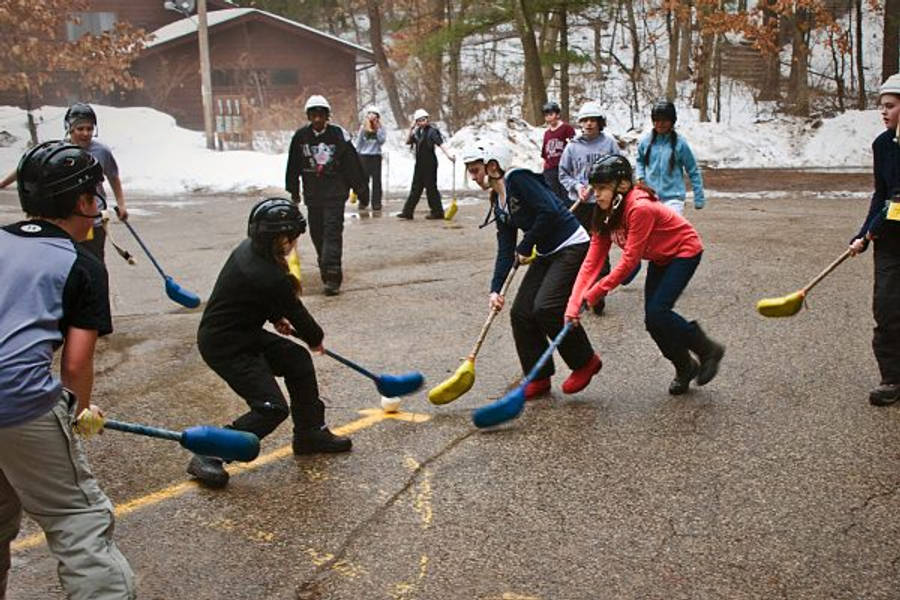 小孩動圖 桌布 壁紙 — 戴著頭盔的孩子們在積雪的瀝青球場上玩街頭曲棍球，背景有被雪覆蓋的樹木和小木屋。
