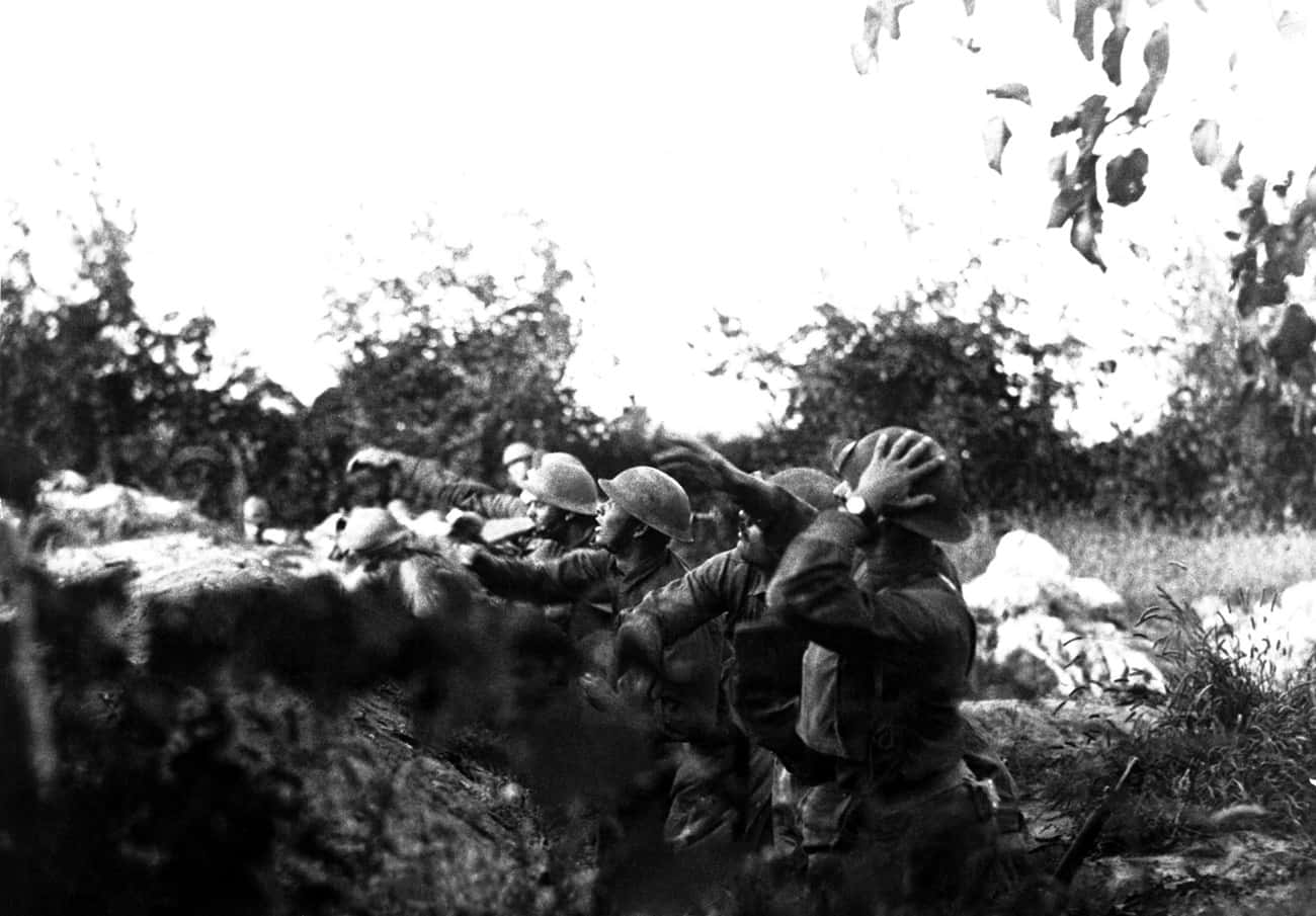 戰爭動圖 桌布 壁紙 — 戴頭盔的士兵在戰壕後方掩護，樹葉和岩石框出嚴酷的戰場場景。