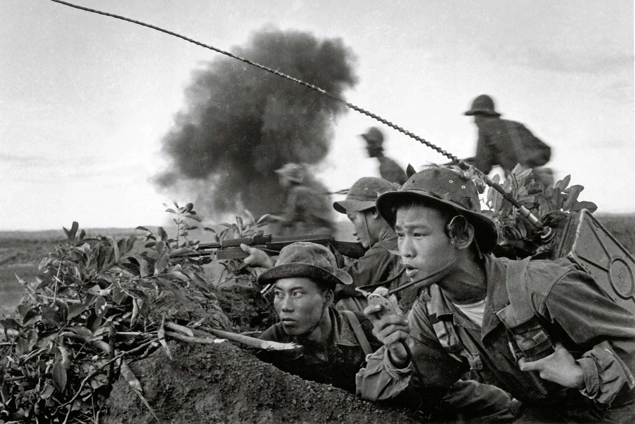 Vietnam War 桌布 壁紙 — 戴頭盔的士兵蹲在泥濘的戰壕中開火，身後煙霧瀰漫的戰場。
