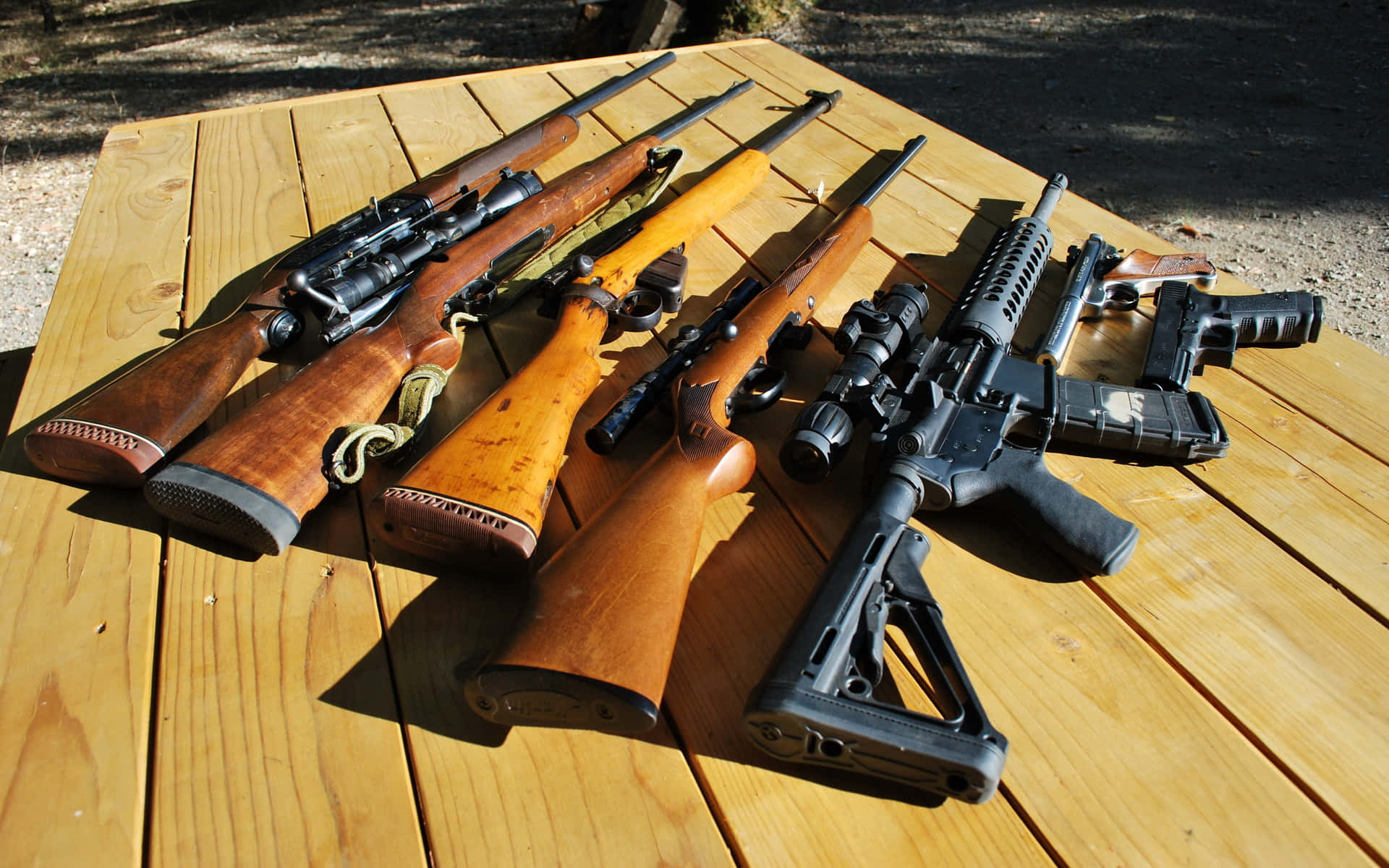 Rifle 桌布 壁紙 — 戶外一張經風化的木製野餐桌上，擺放著陽光照射下的步槍和手槍收藏。