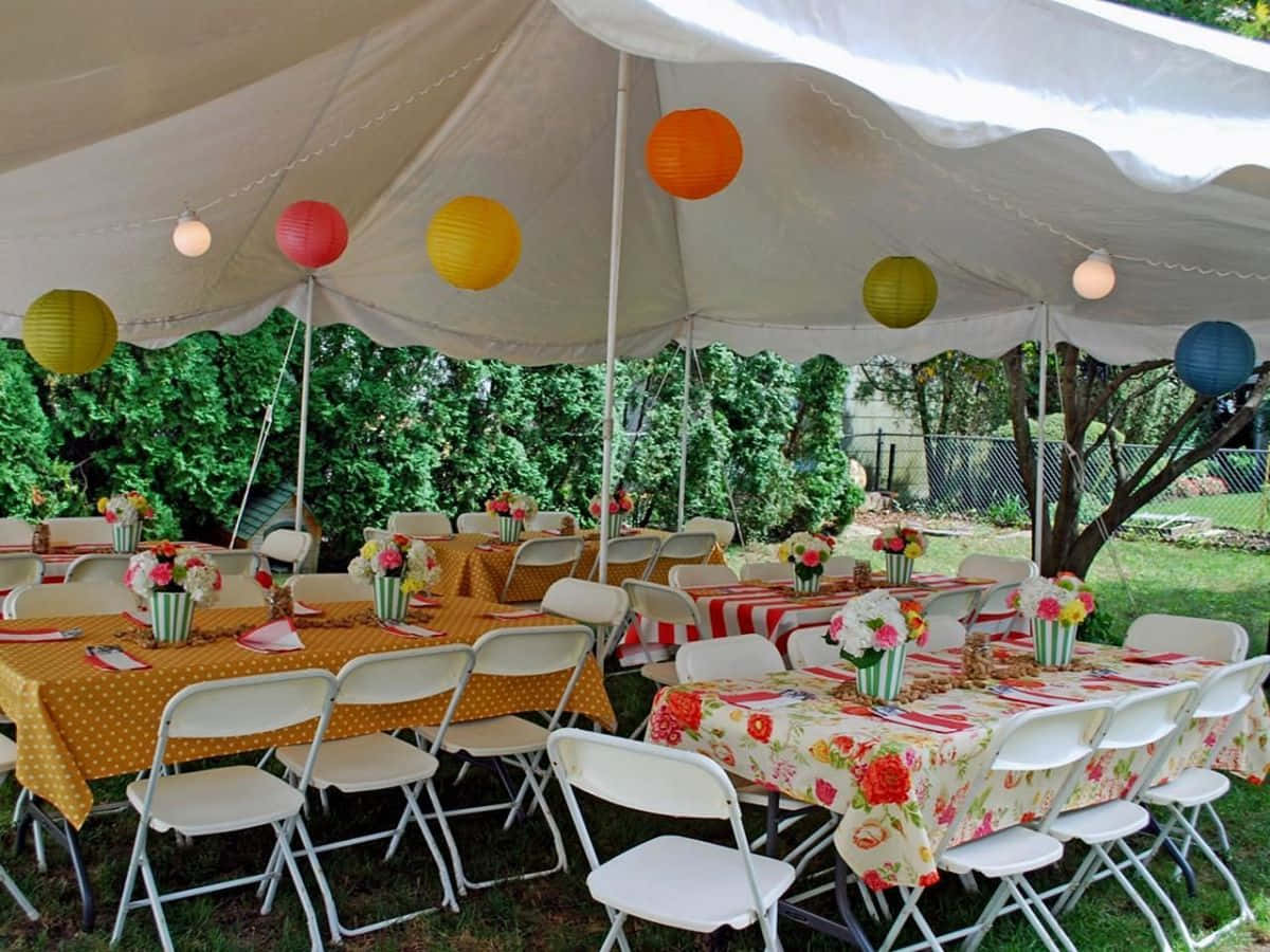 Birthday Party 桌布 壁紙 — 戶外派對餐桌在白色帳篷下，裝飾著彩色紙燈籠和花卉桌布。