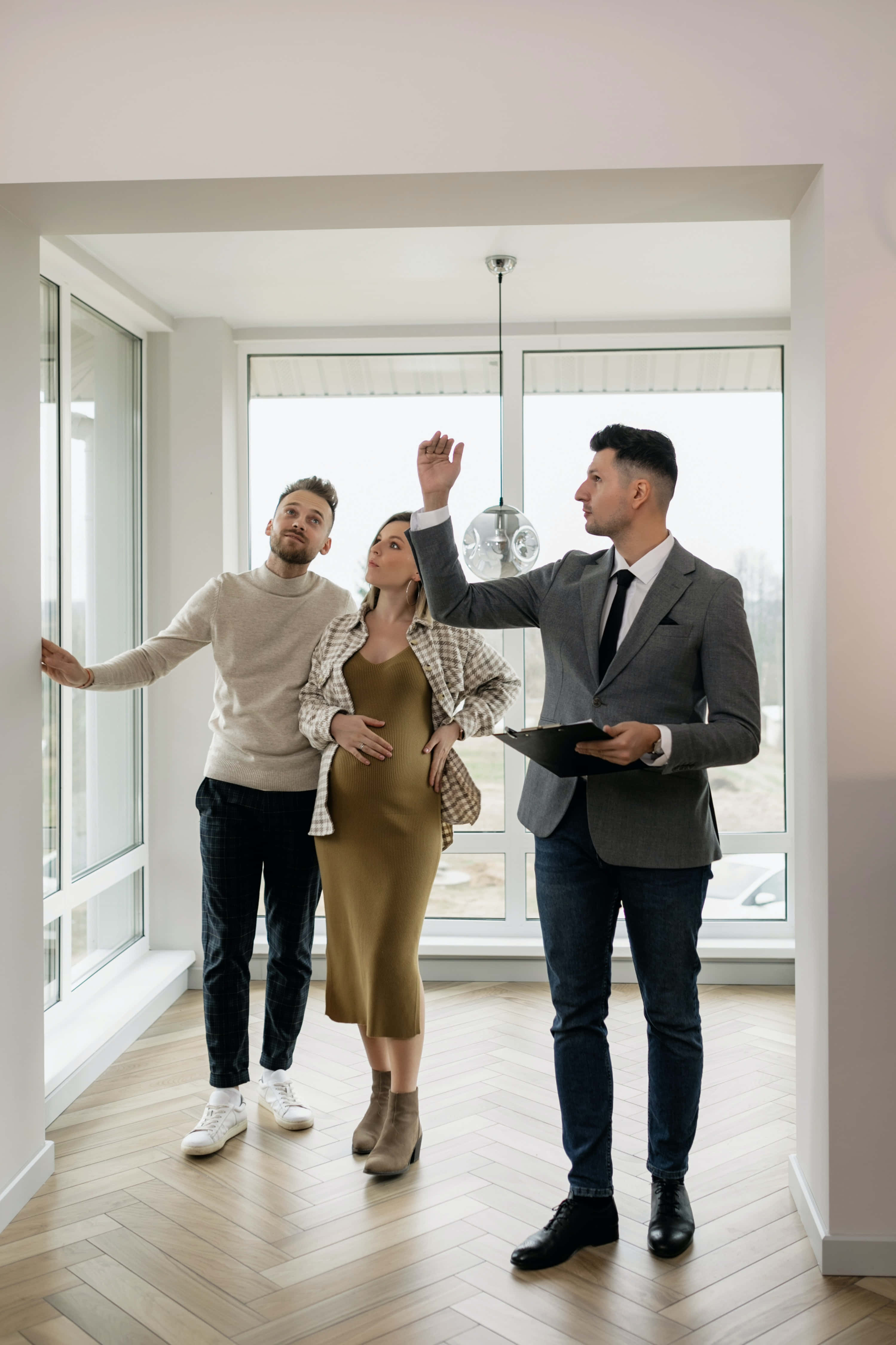 Real Estate Agent 桌布 壁紙 — 房地產經紀人指著天花板燈，而一對懷孕夫婦在明亮現代、鋪有人字紋木地板的公寓中觀看。
