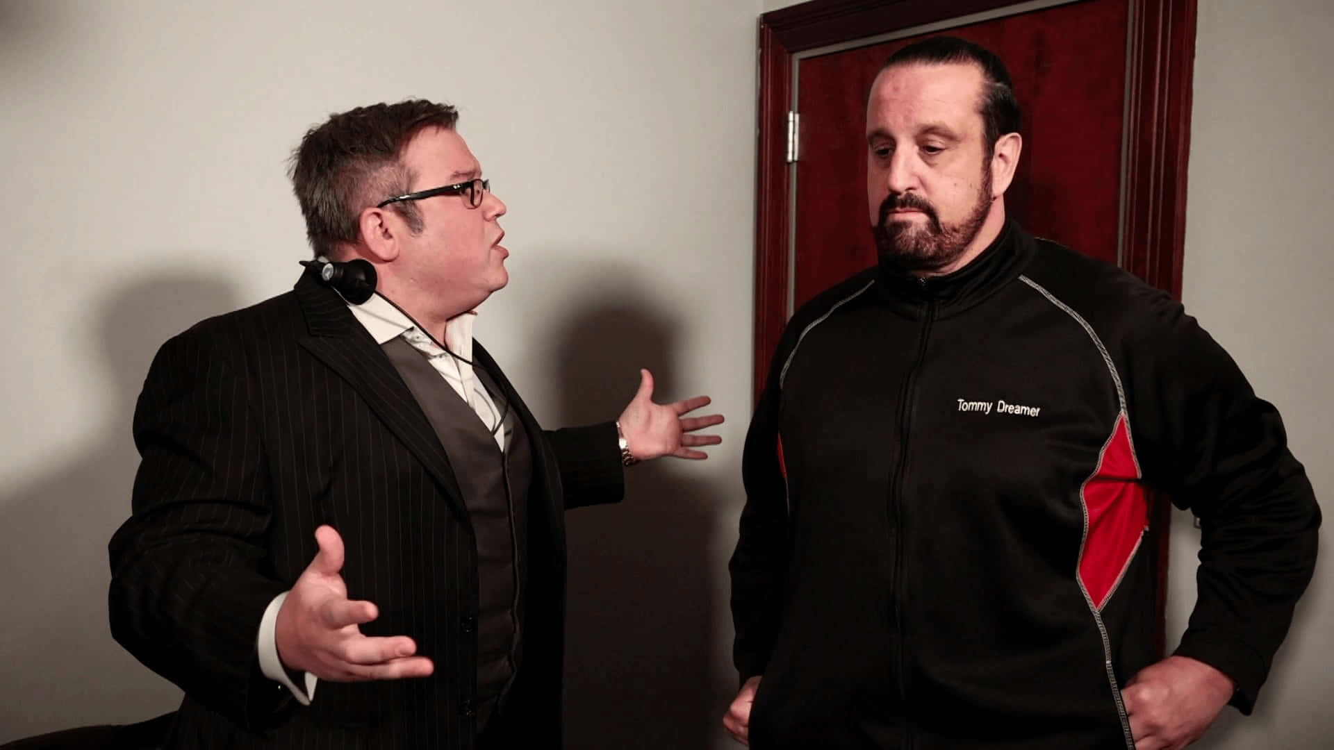 WWE 桌布 壁紙 — 房間裡有兩個男人，一個身穿西裝生動地做手勢，另一個穿著帶紅色點綴的黑色外套專心聆聽。
