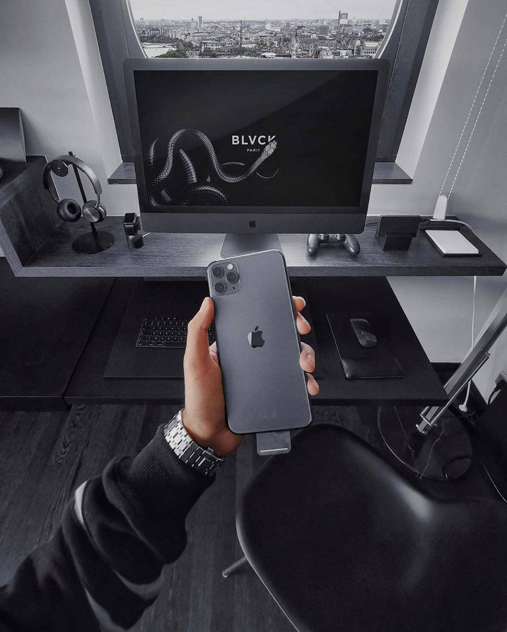 iPhone 桌布 壁紙 — 手在時尚黑色辦公桌前拿著深灰色iPhone，顯示器顯示蛇標誌和透過窗戶的城市景觀。