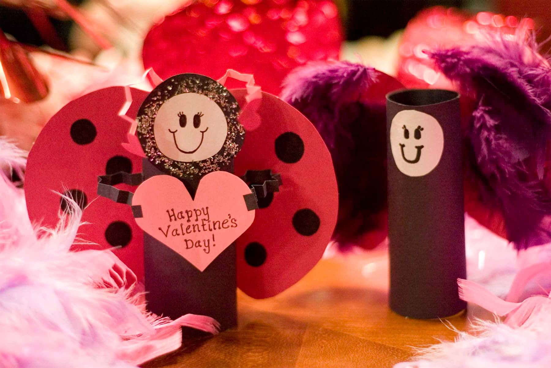 Cute Happy Valentine Day 桌布 壁紙 — 手工情人節瓢蟲工藝品，帶有閃閃發光的笑臉和心形，上面寫著「Happy Valentine's Day!」，周圍環繞著粉紅色和紫色的羽毛。