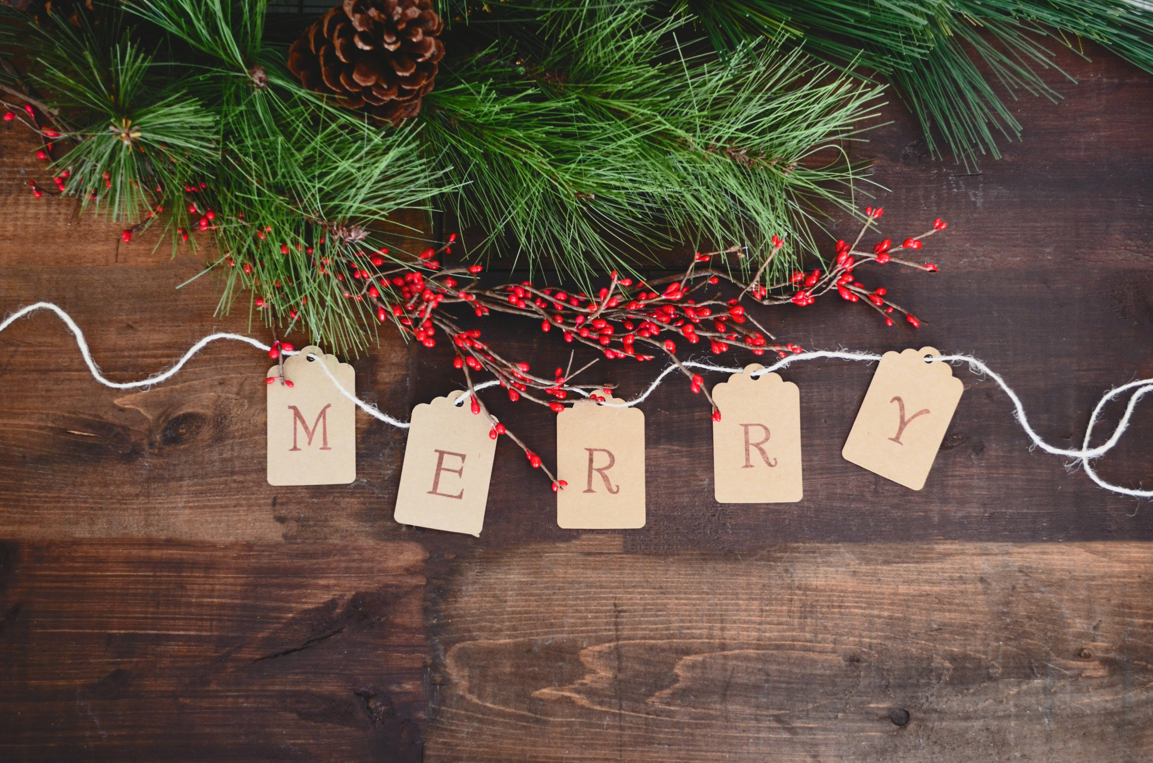 Greetings 桌布 壁紙 — 手工製作的 'MERRY' 標籤用麻繩和紅色漿果串起，放在質樸的木頭上，旁邊有松針和松果。