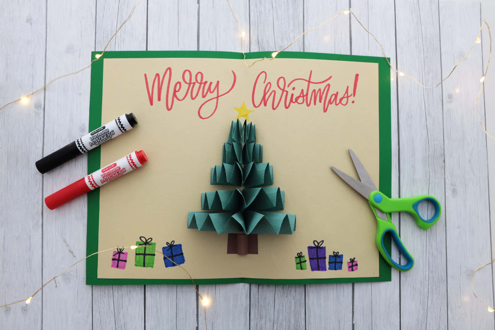 Christmas Card 桌布 壁紙 — 手工製作的3D紙聖誕樹，附有禮物和寫著「聖誕快樂」的卡片，周圍環繞著手工藝用品和仙女燈。