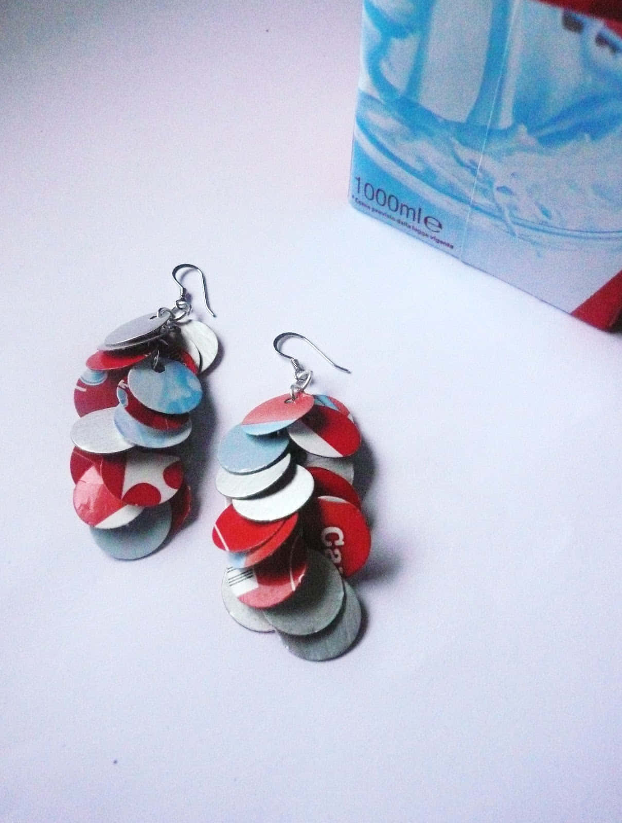 Earrings 桌布 壁紙 — 手工製作的耳環，由堆疊的紅色、白色和藍色圓形汽水罐拉環製成，掛在一個1000毫升水瓶盒旁邊的純白表面上。