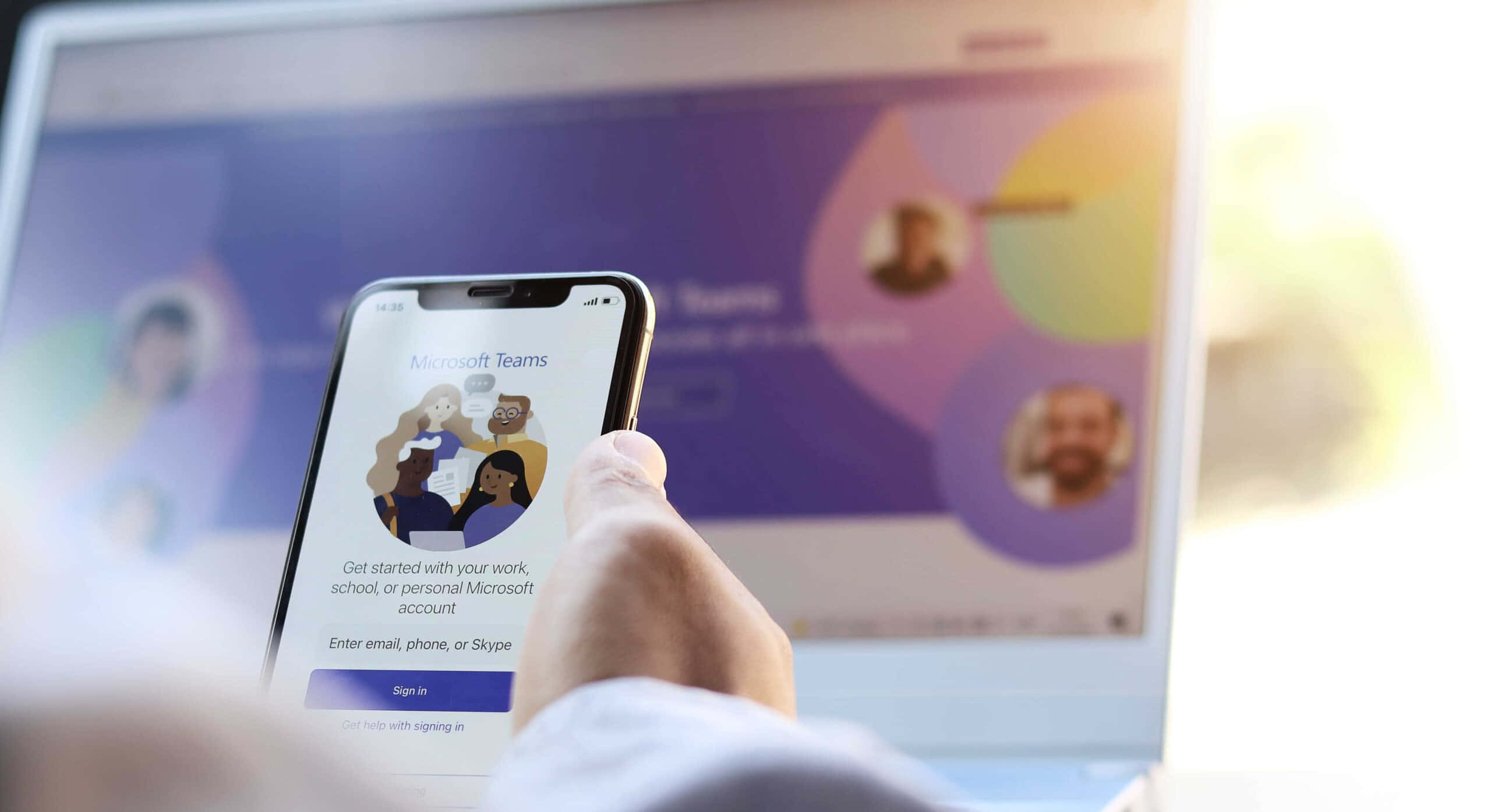Microsoft Teams 桌布 壁紙 — 手拿著顯示Microsoft Teams登入畫面的智慧型手機，背景有模糊的筆記型電腦顯示團隊介面。
