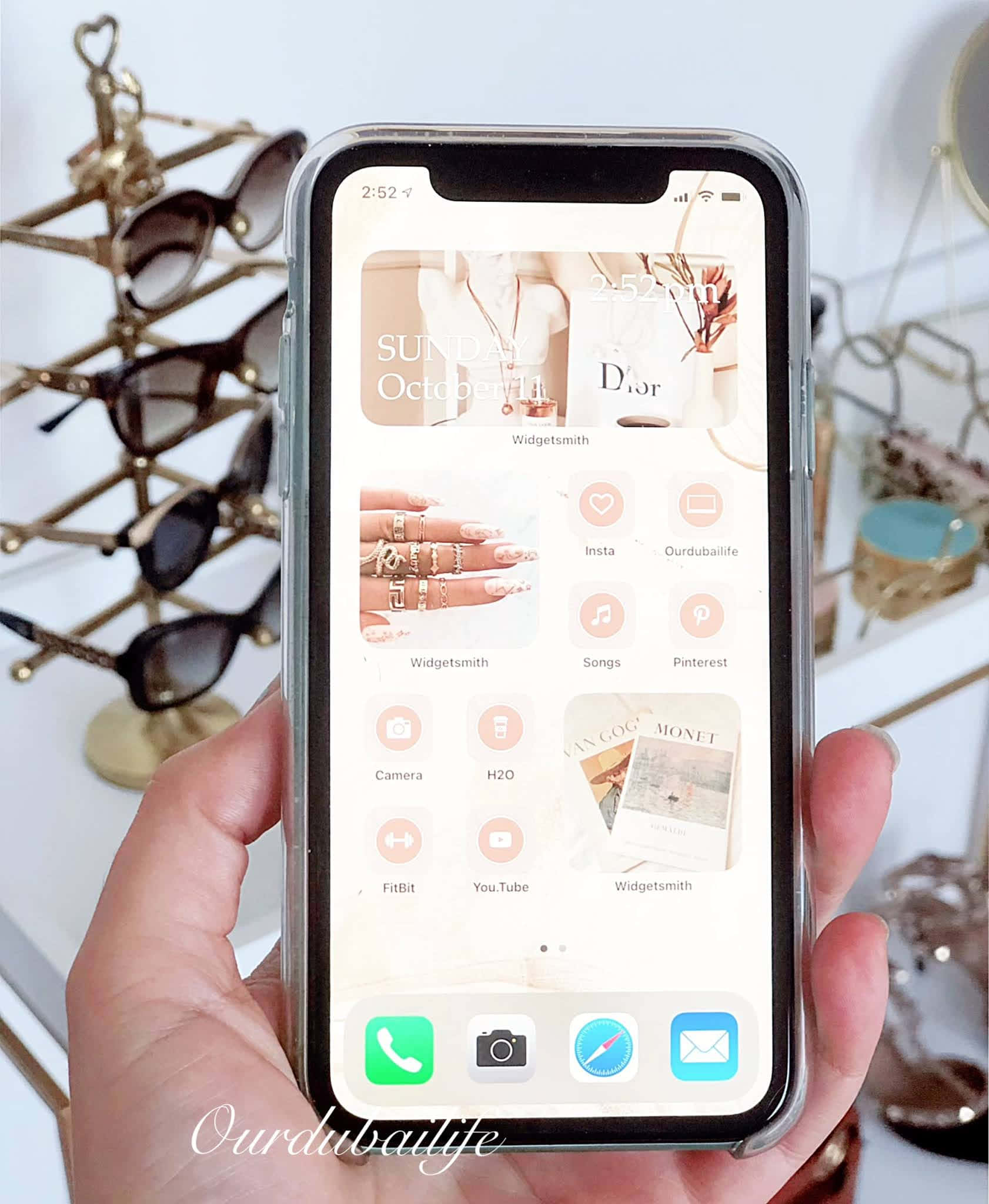 iPhone 美學 桌布 壁紙 — 手握住手機，展示時尚簡約的桌布，帶有時尚小工具和應用程式圖標，背景為金色太陽眼鏡。