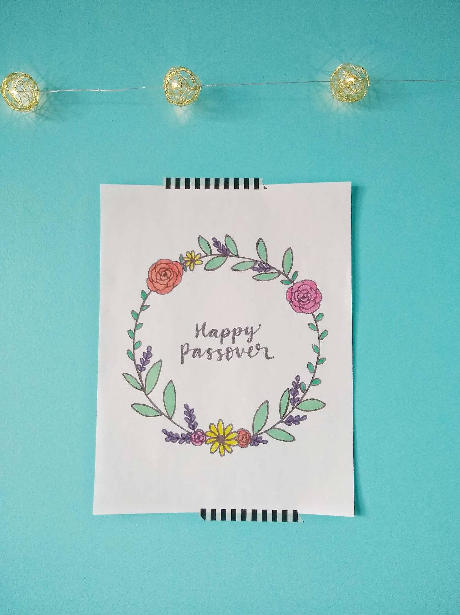 Passover 桌布 壁紙 — 手繪花環，白色記事本上寫有「Happy Passover」字樣，背景為藍綠色牆壁及燈串。