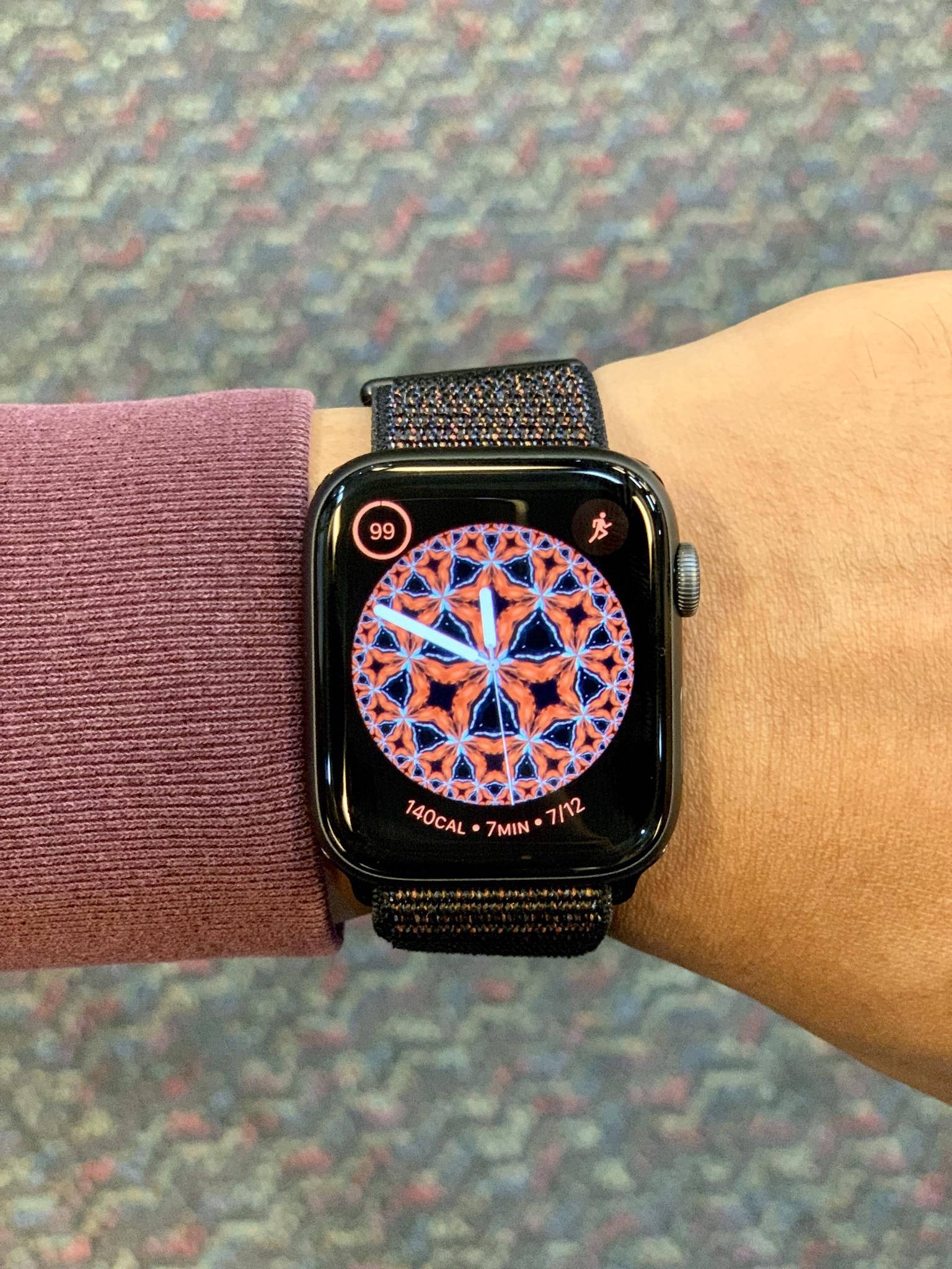 Apple Watch 動態 桌布 壁紙 — 手腕上智慧手錶的特寫，在圖案地毯背景下顯示充滿活力的幾何鍛鍊錶盤。