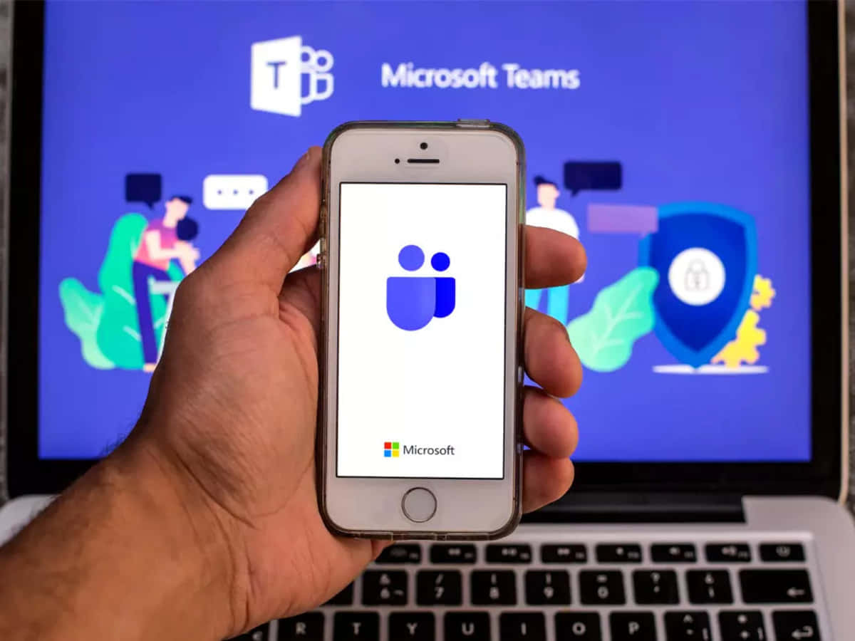 Microsoft Teams 桌布 壁紙 — 手裡拿著智慧型手機上的 Microsoft Teams 應用程式，前方筆記型電腦顯示應用程式的插圖介面，包含人物和盾牌圖示。