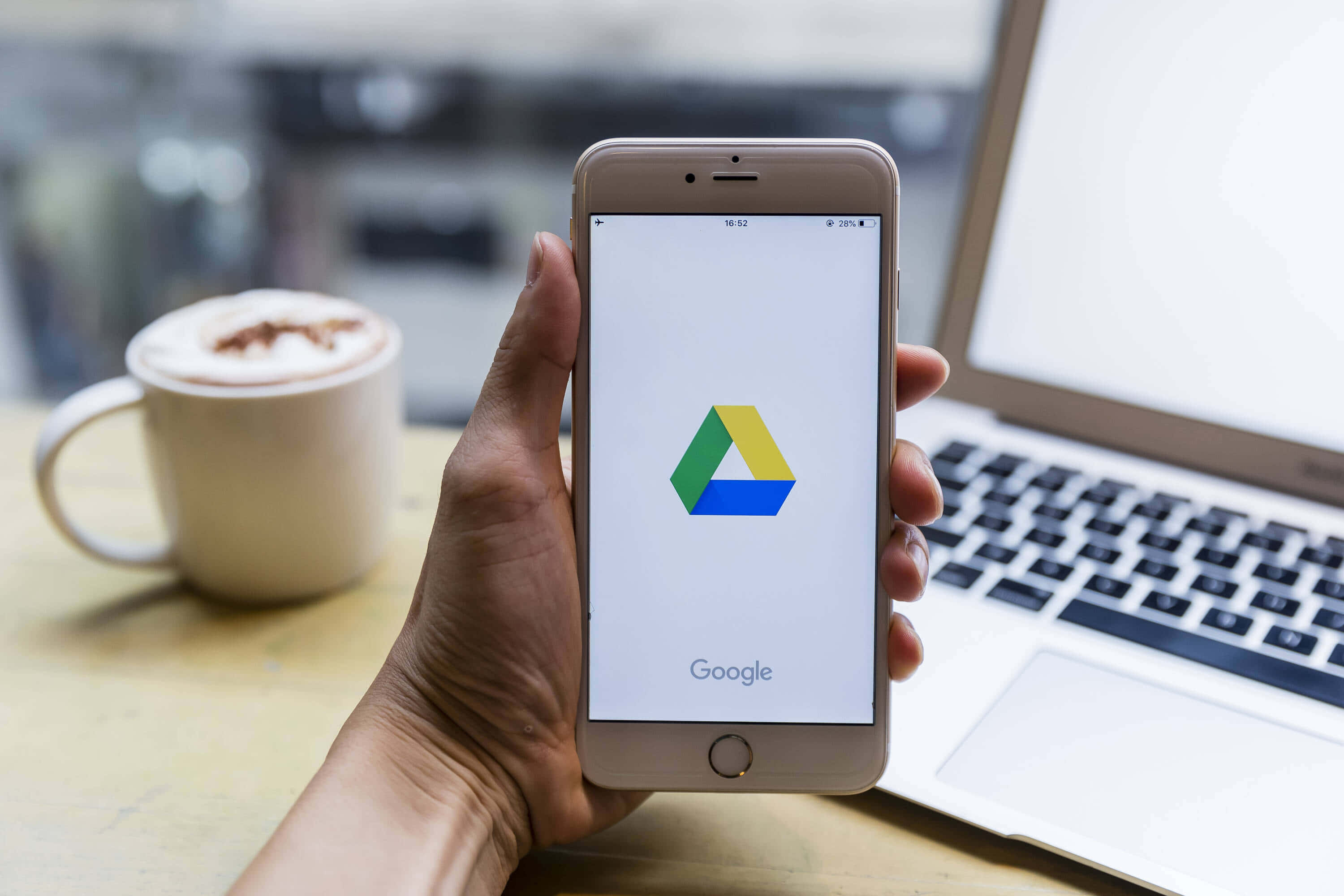 科技動圖 桌布 壁紙 — 手裡拿著顯示 Google Drive 標誌的智慧型手機，旁邊有咖啡杯和筆記型電腦在木桌上。