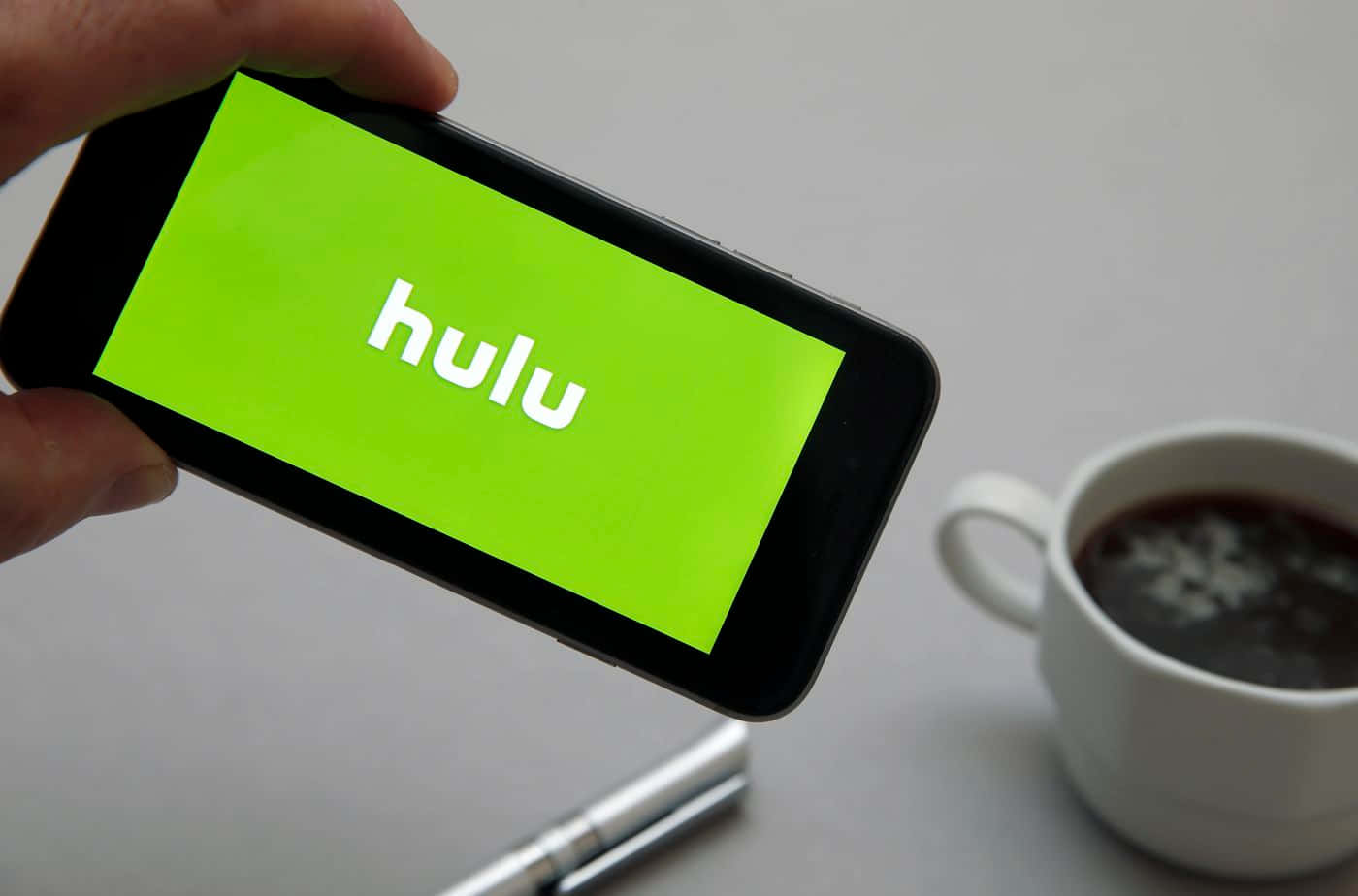 Hulu 桌布 壁紙 — 手裡拿著顯示Hulu標誌的智慧型手機，旁邊放著一杯咖啡，在簡約風格的桌子上。