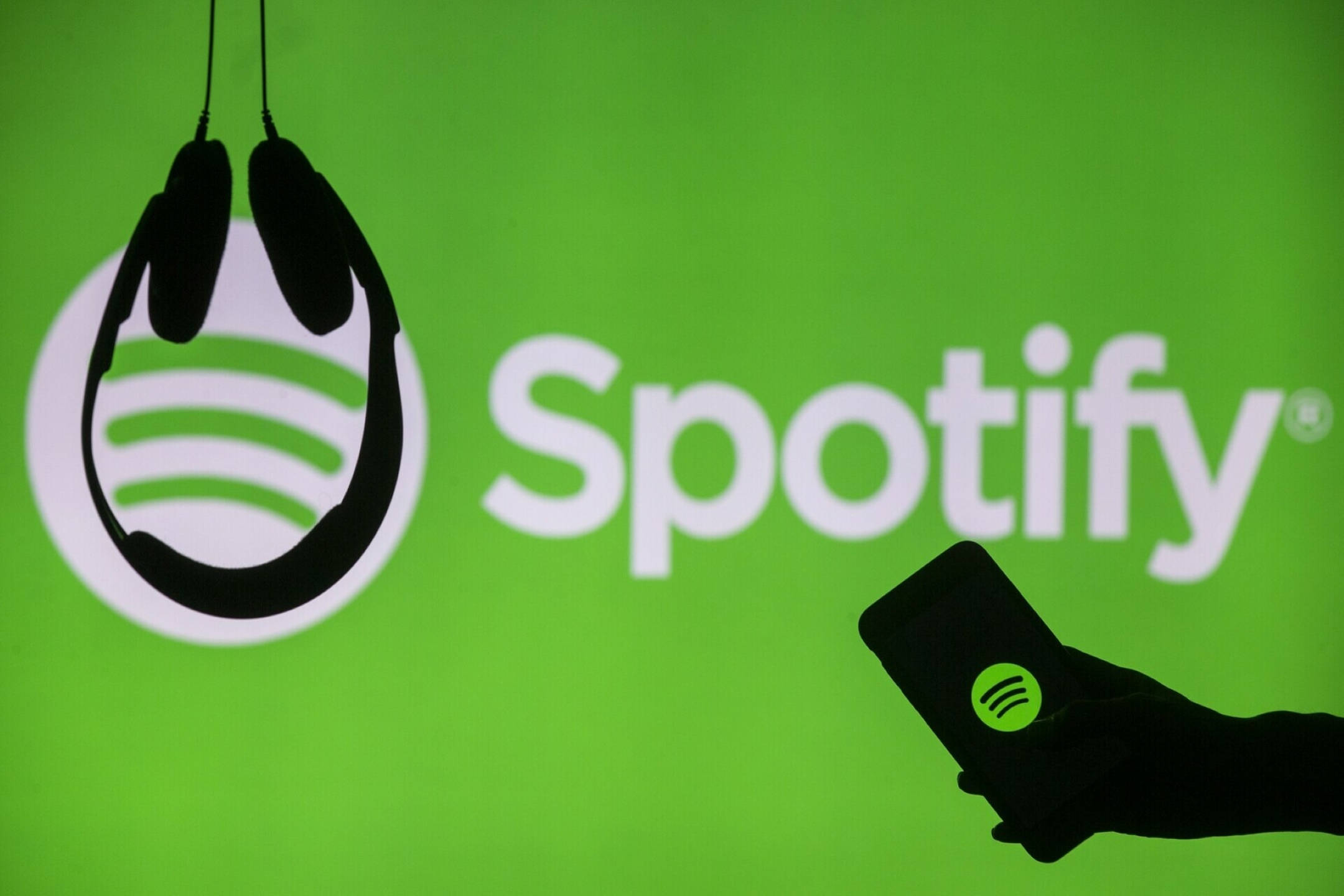 Spotify 桌布 壁紙 — 手部剪影握著帶有Spotify標誌的手機，背景為明亮的綠色品牌背景，並有懸掛的耳機。