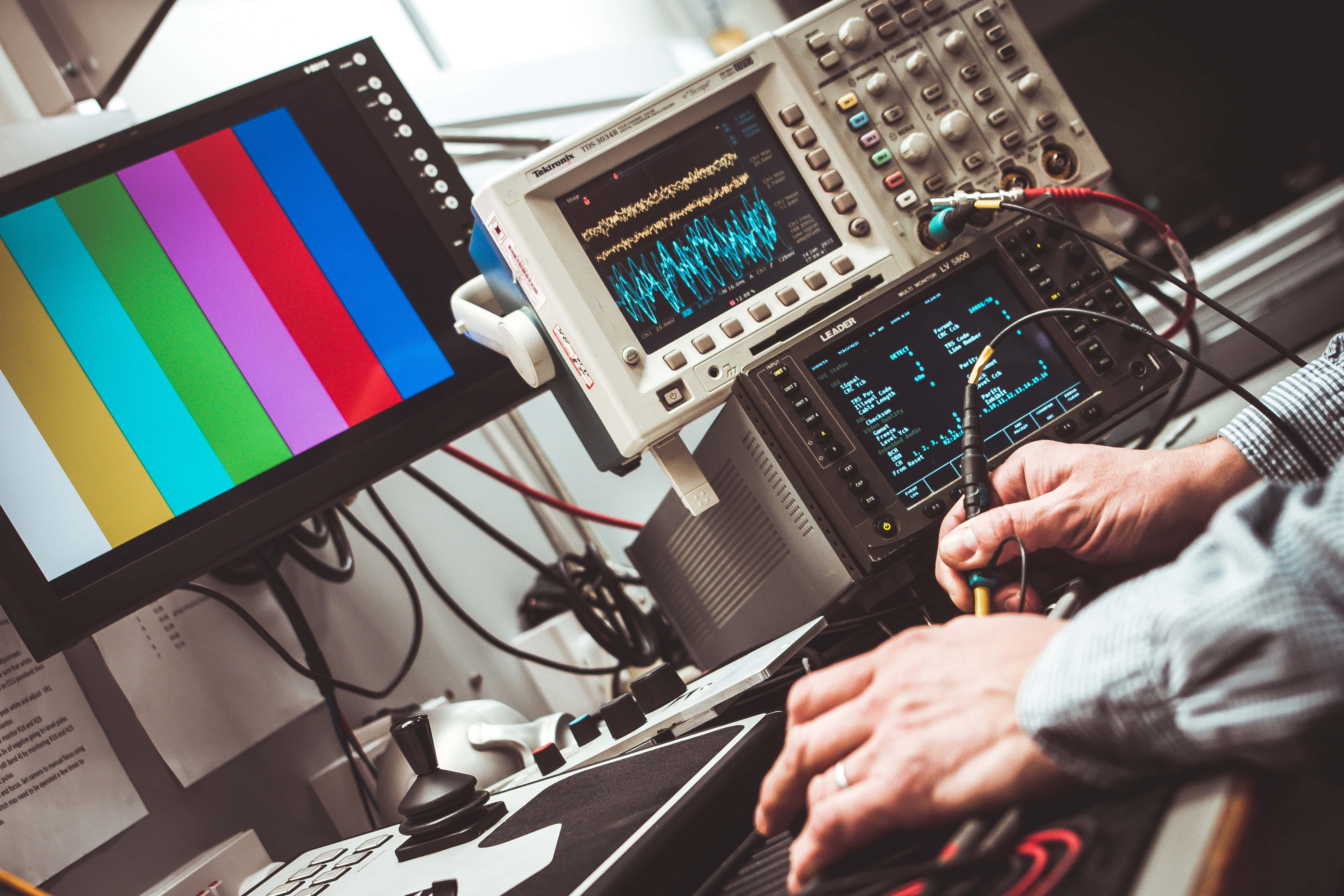 Monitor 桌布 壁紙 — 技術人員的手在雜亂的廣播實驗室中，調整示波器上的探頭，旁邊是彩色條測試監視器。