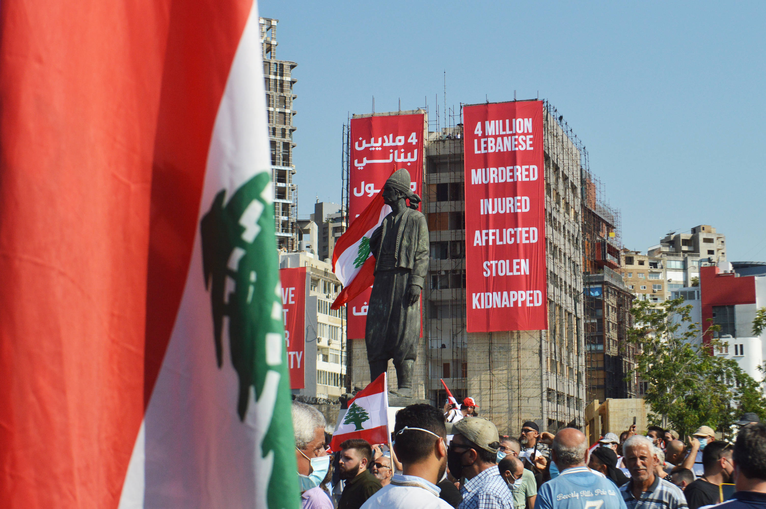 Lebanon 桌布 壁紙 — 抗議者聚集在陽光明媚的城市廣場，在雕像附近揮舞黎巴嫩國旗和標語，標語上寫著「400萬黎巴嫩人被殺害、受傷、受苦、被盜、被綁架」。