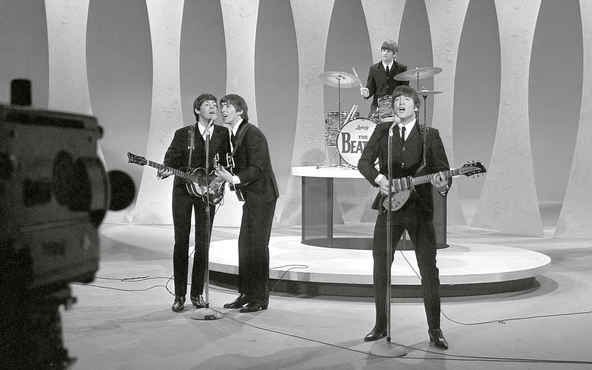 the beatles 桌布 壁紙 — 披頭四在一個帶有弧形背景和印有他們名字的黑白鼓組的復古舞台上表演。