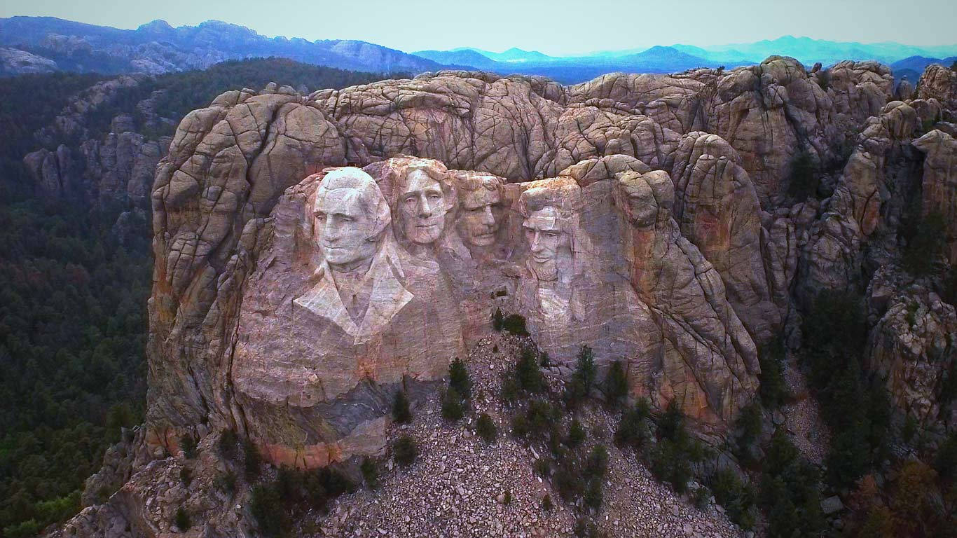 Mount Rushmore 桌布 壁紙 — 拉什莫爾山雕刻的總統面孔的鳥瞰圖，位於崎嶇的花崗岩懸崖上，周圍環繞著森林覆蓋的山脈。