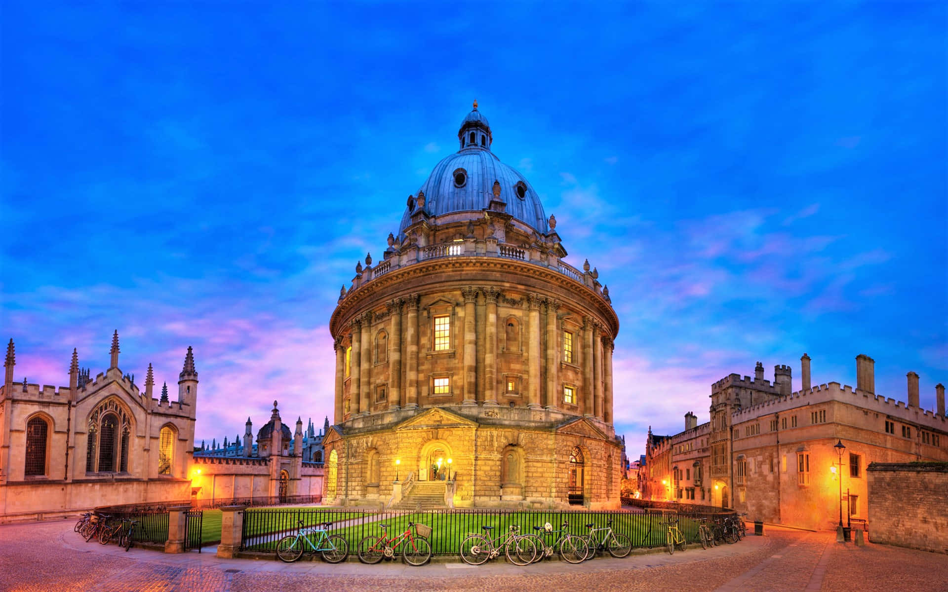 England 桌布 壁紙 — 拉德克利夫相機樓沐浴在金黃色的暮光中，映襯著充滿活力的藍色和紫色天空，附近停著自行車。