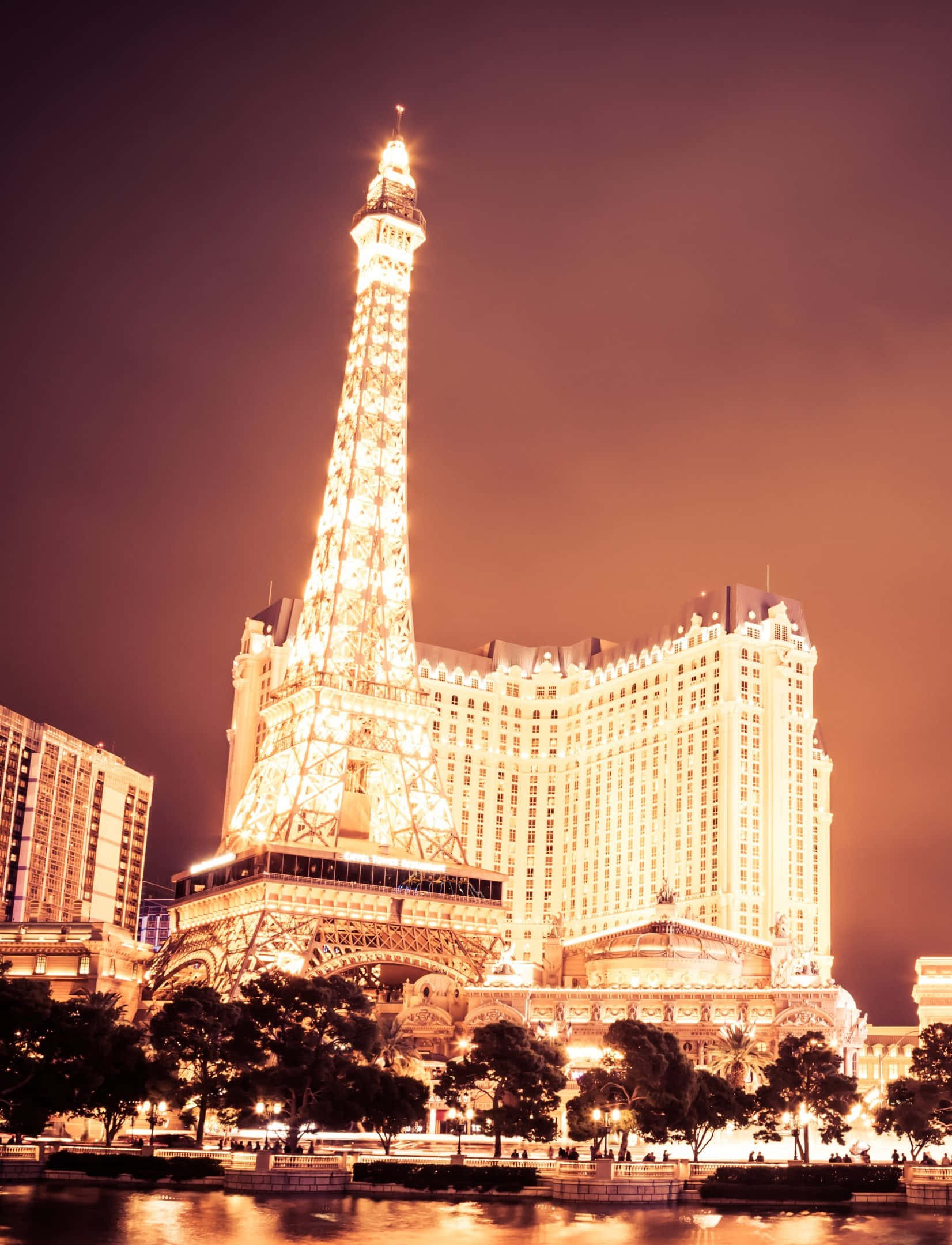 Las Vegas Phone 桌布 壁紙 — 拉斯維加斯一間大酒店頂上，一座耀眼的艾菲爾鐵塔複製品在夜間溫暖發光，倒映在下方平靜的水道中。