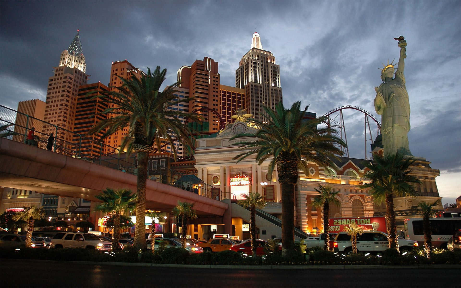 Las Vegas Skyline 桌布 壁紙 — 拉斯維加斯充滿活力的黃昏場景， featuring 自由女神像複製品、棕櫚樹和戲劇性多雲天空下閃閃發光的賭場建築。