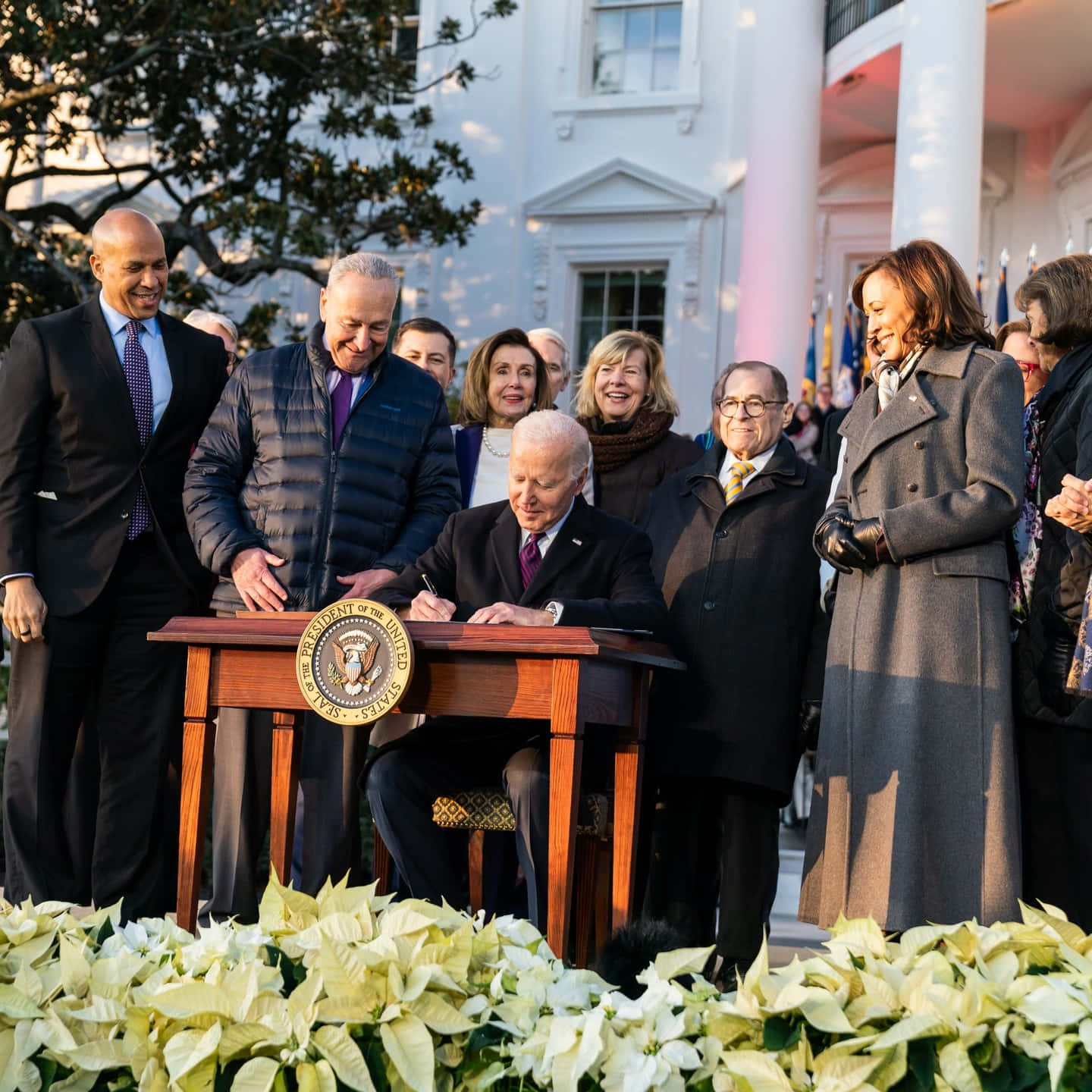 Kamala Harris 桌布 壁紙 — 拜登總統在木製講台上簽署法案，微笑的官員和賓客在戶外聚集在他周圍。