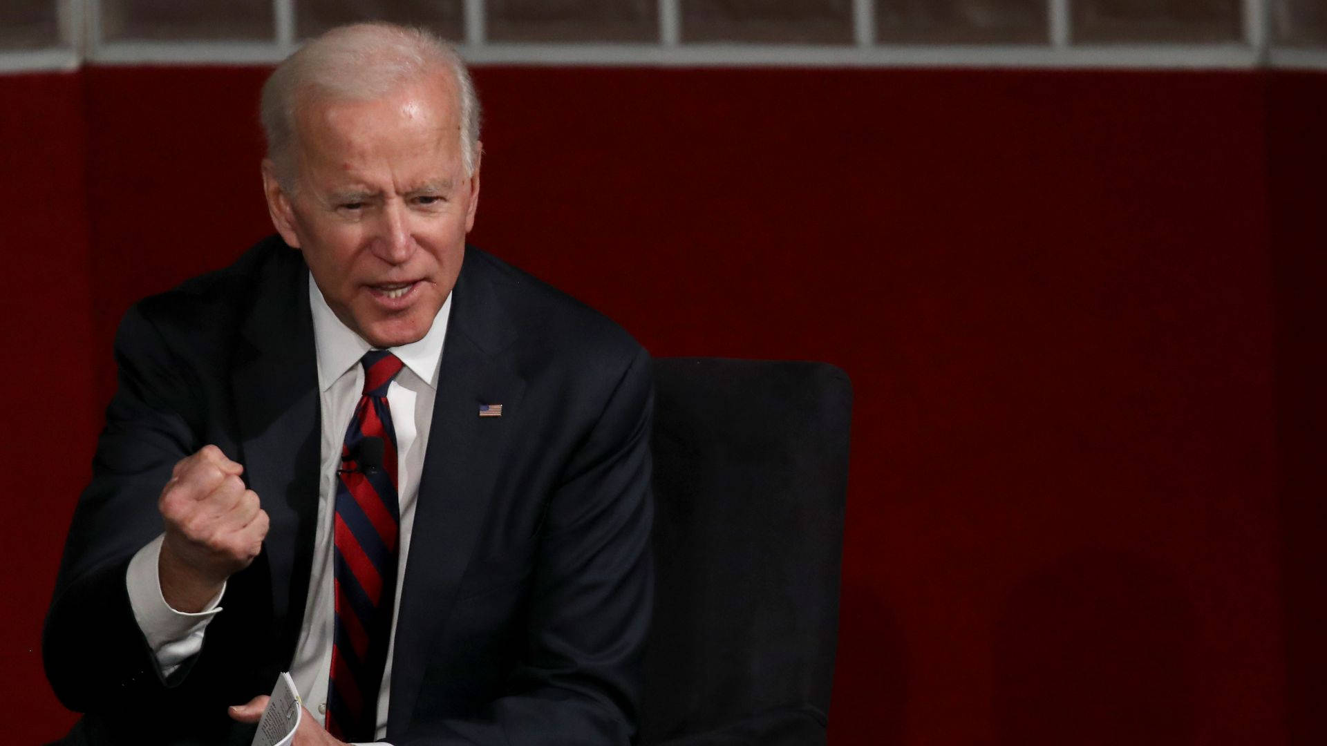 Biden 桌布 壁紙 — 拜登總統坐在紅色背景前的椅子上，說話時緊握拳頭，強調手勢。