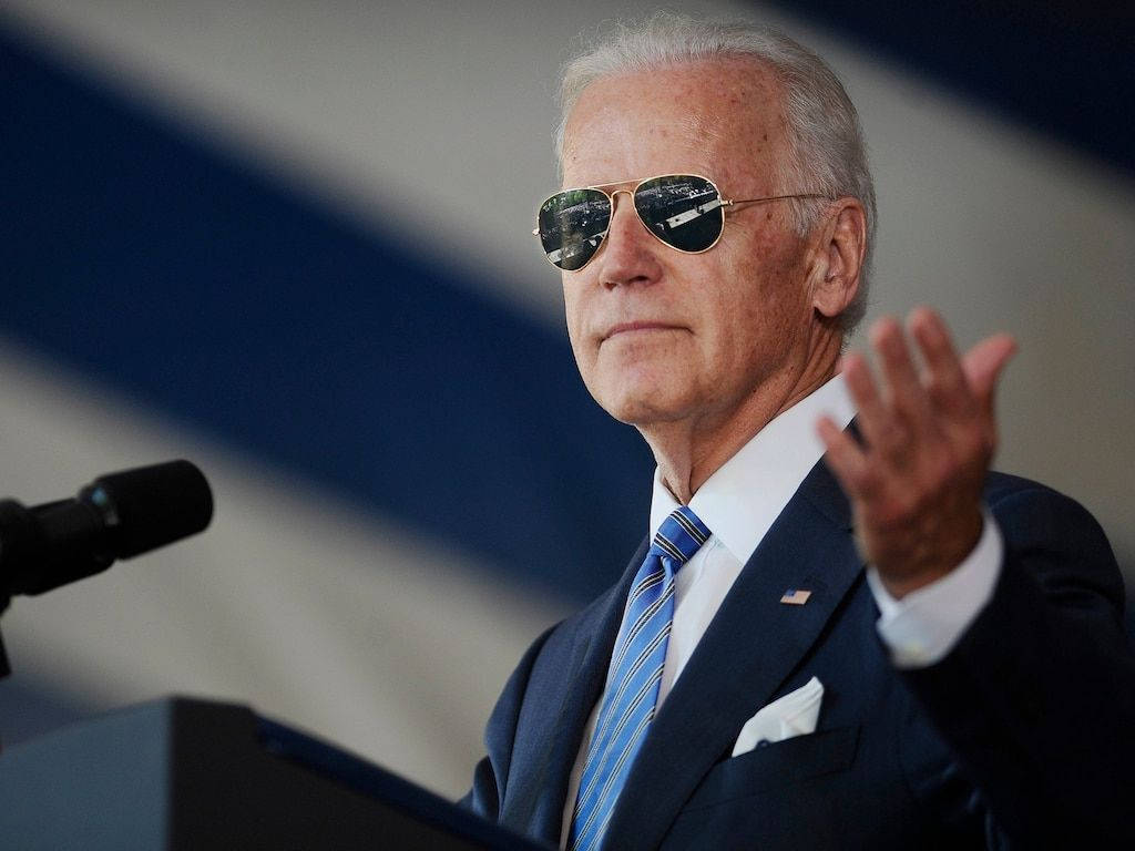 Biden 桌布 壁紙 — 拜登總統戴著飛行員太陽眼鏡在講台上揮手，背景是模糊的旗幟。