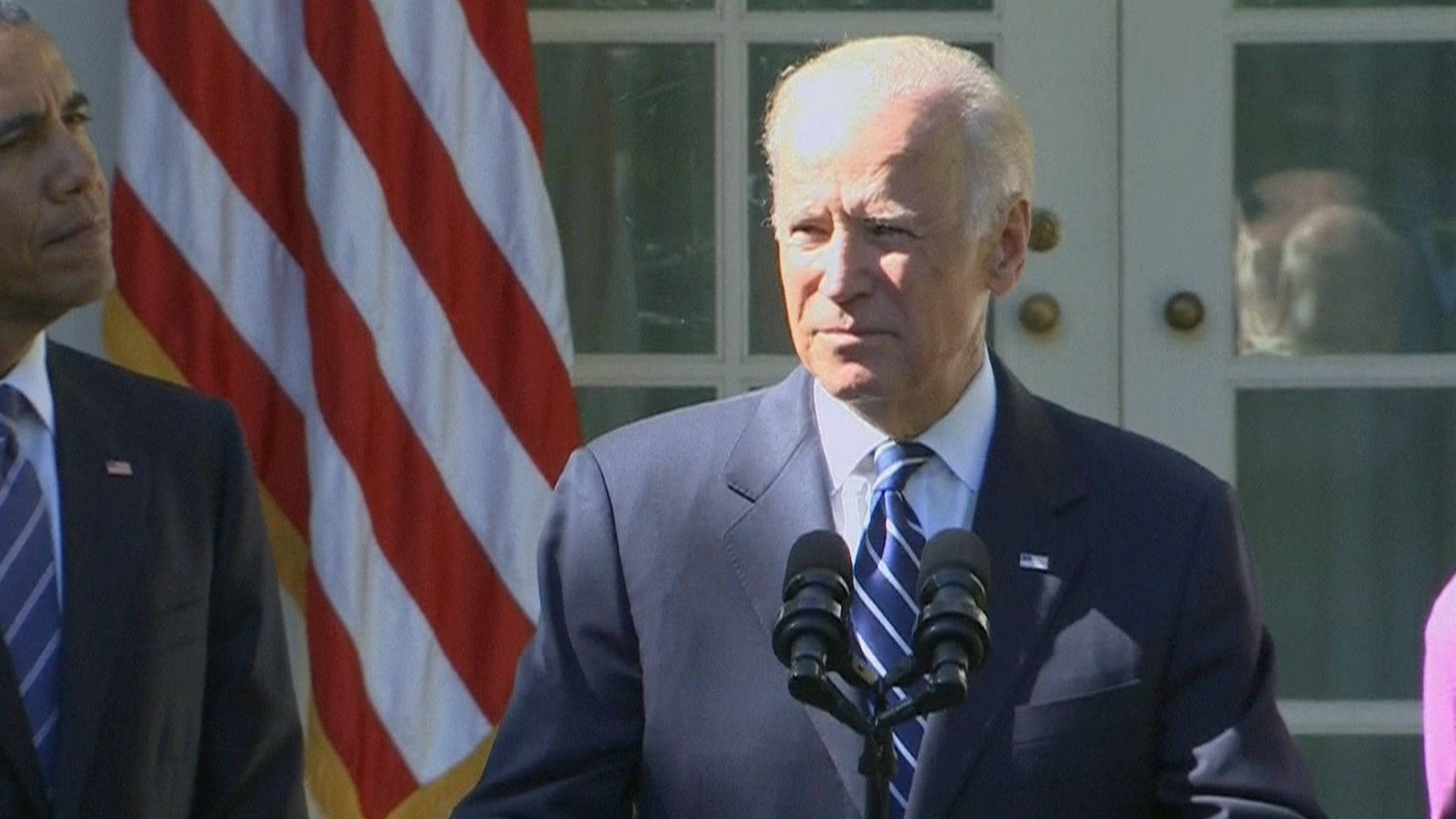 Biden 桌布 壁紙 — 拜登總統站在講台上，前面有麥克風，背景是美國國旗和白色門，在正式戶外記者會期間。