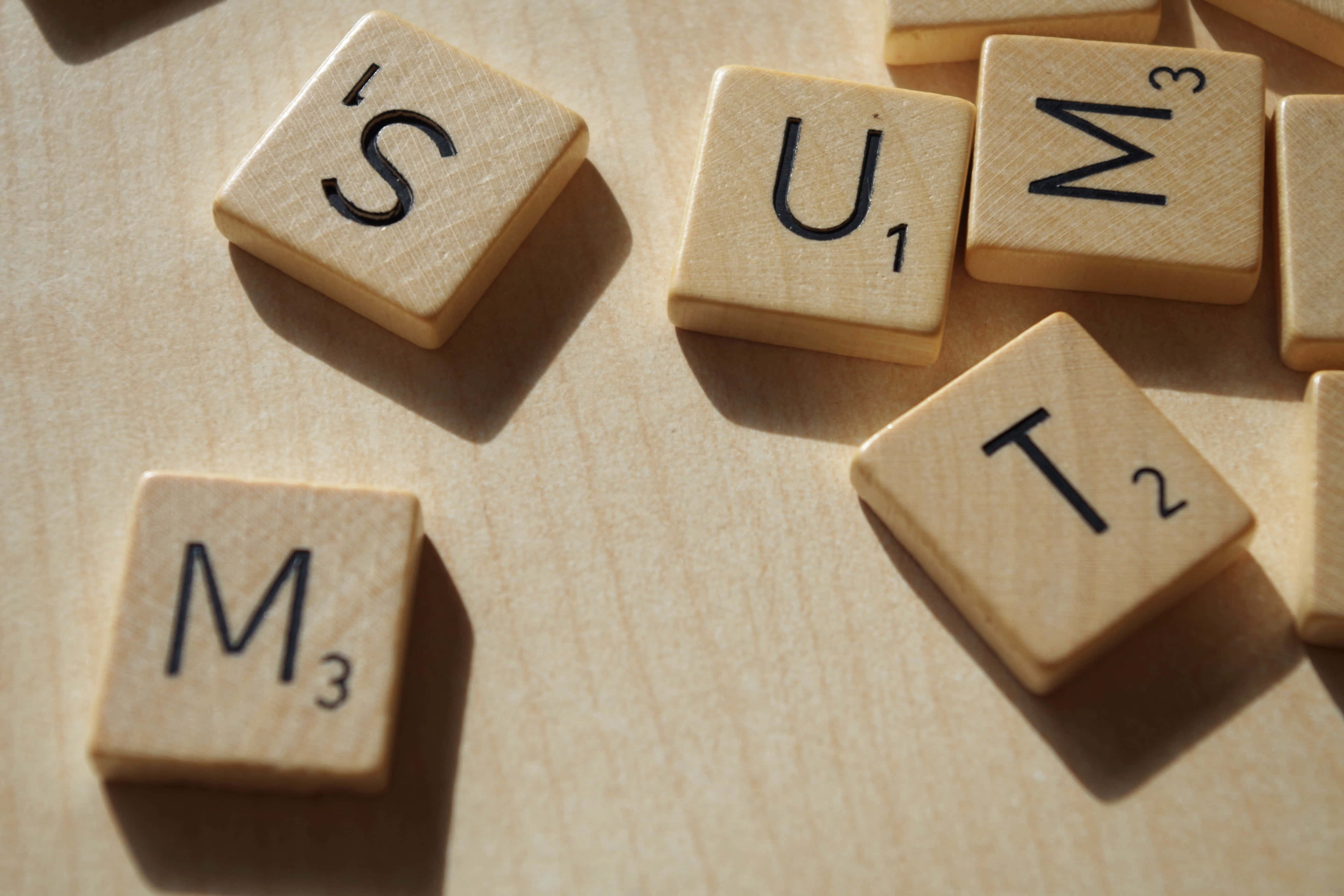 Educational Games 桌布 壁紙 — 拼出「SUM」和「IS」的 Scrabble 字母牌放在淺色木質表面上，帶有柔和陰影。