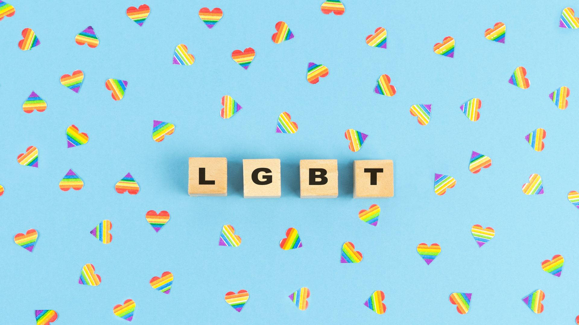 彩虹 桌布 壁紙 — 拼出LGBT的木塊在淺藍色表面上佔據中心位置，周圍散落著彩虹心形紙屑。