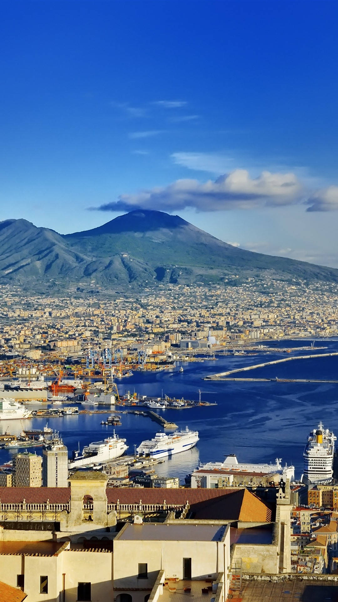 Naples 桌布 壁紙 — 拿坡里的全景景觀，維蘇威火山聳立於城市和擠滿郵輪的港口之上，天空清澈湛藍。