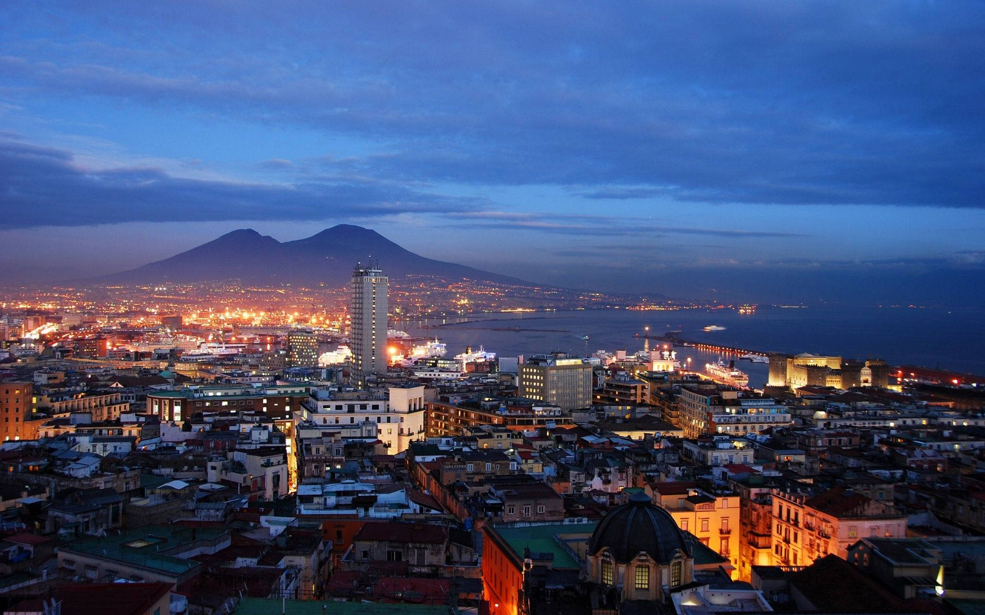Naples 桌布 壁紙 — 拿坡里黃昏城市景觀，閃爍燈光、海灣和維蘇威火山在深藍色天空的襯托下形成剪影。