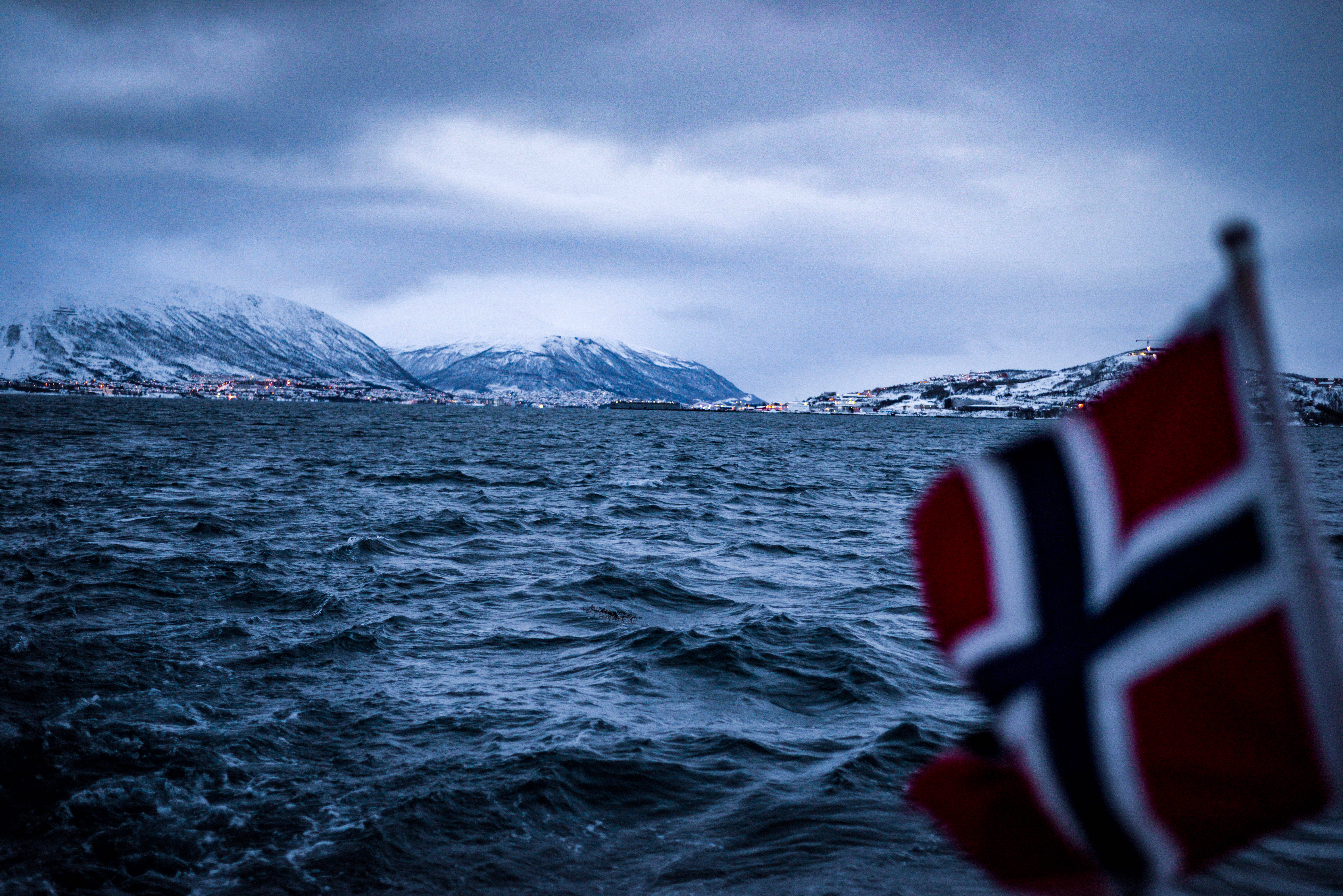 Norway 桌布 壁紙 — 挪威國旗在前景飄揚，而一個白雪覆蓋的山城在多雲的黃昏天空下閃耀。