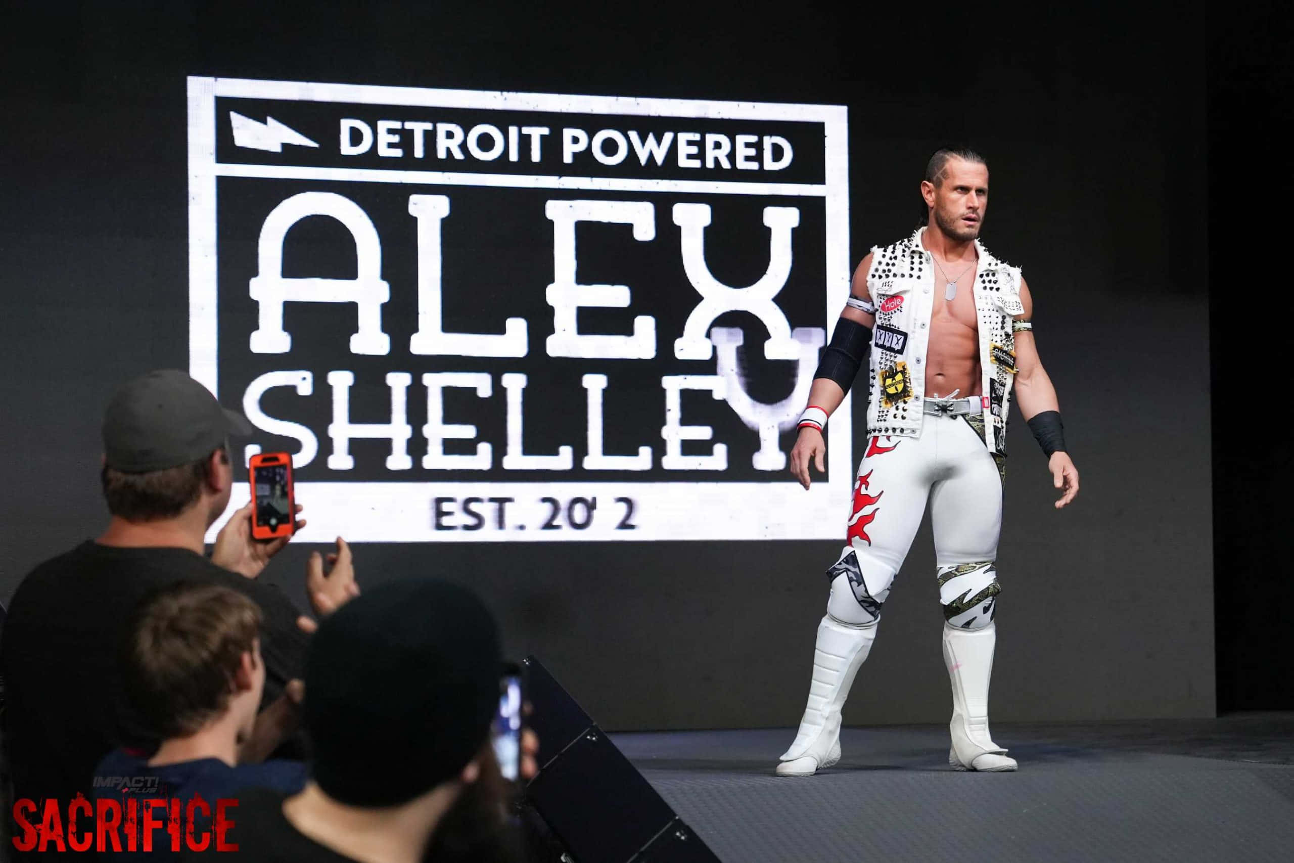 摔角 桌布 壁紙 — 摔角手 Alex Shelley 身穿白色裝備，自信地站在舞台上，身後是巨大的「Detroit Powered」標誌，粉絲們正在記錄這一刻。