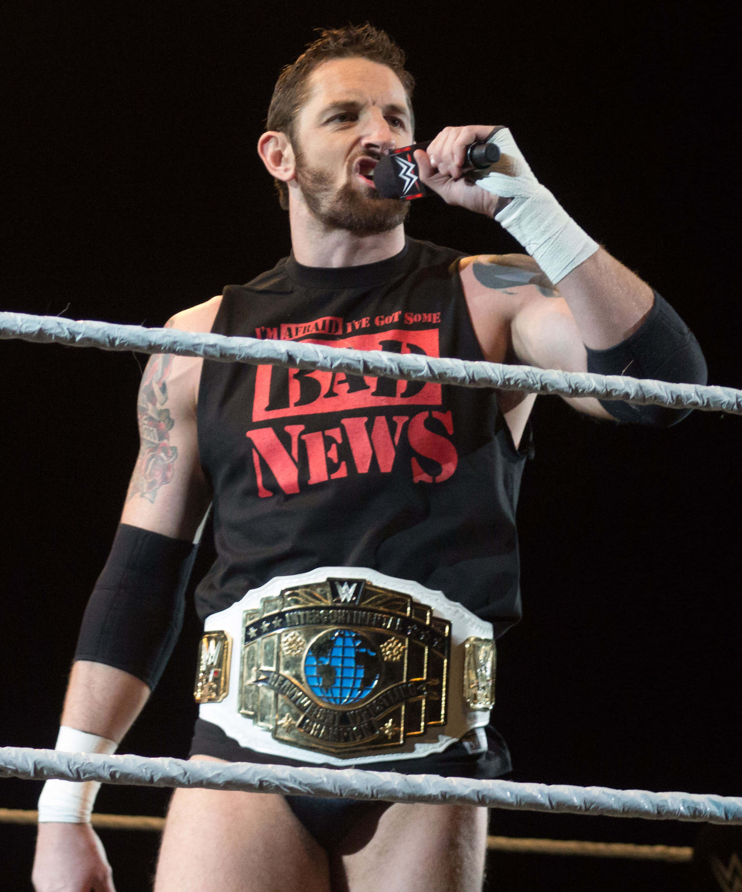 WWE 桌布 壁紙 — 摔角手 Bad News Barrett 在擂台上手持麥克風，戴著冠軍腰帶和印有紅色文字的黑色背心。