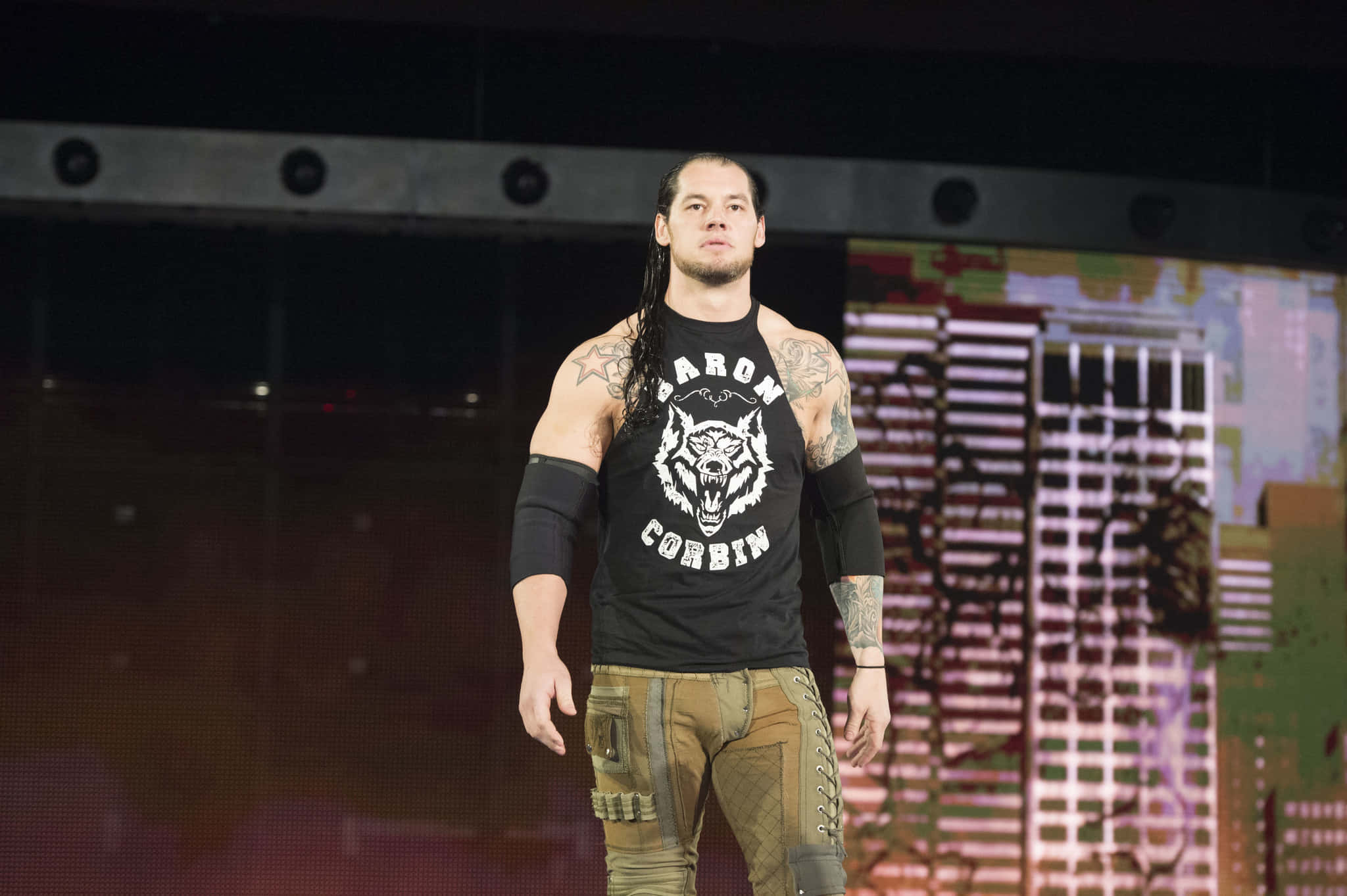 WWE 桌布 壁紙 — 摔角手 Baron Corbin 在場館燈光下自信地站在舞台上，身穿黑色 T 恤和工裝褲。