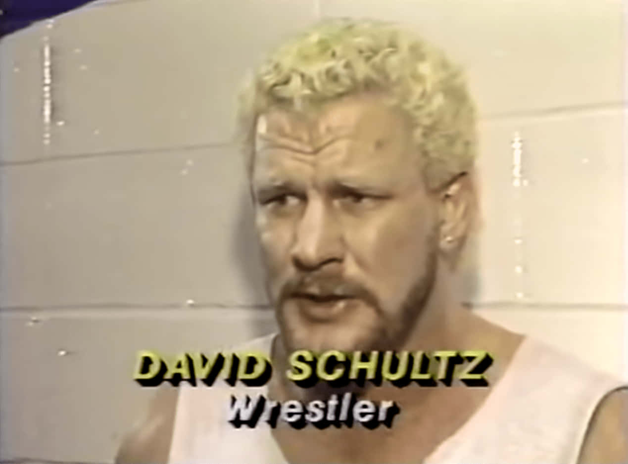 WWE 桌布 壁紙 — 摔角手 David Schultz 的近照，金色捲髮和鬍鬚，身穿白色背心，背景是瓷磚牆。