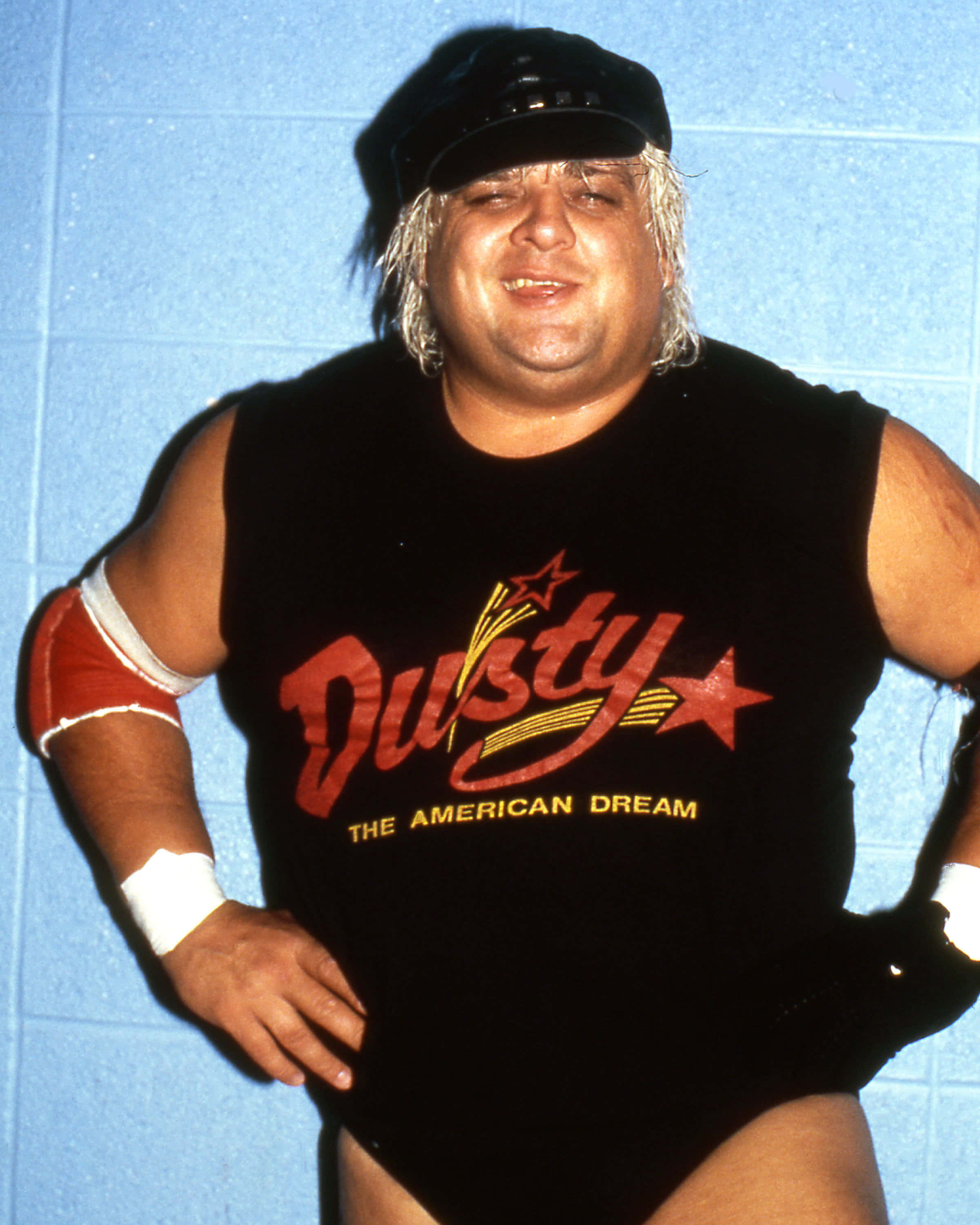 Dusty Rhodes 桌布 壁紙 — 摔角手 Dusty Rhodes 身穿標誌性黑色緊身衣，帶有紅色星星標誌，在藍色煤渣磚牆前自信地擺姿勢。