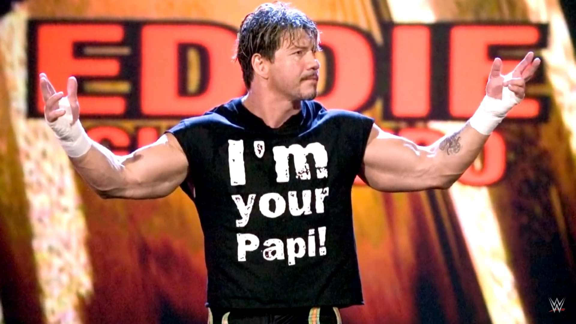 摔角 桌布 壁紙 — 摔角手 Eddie Guerrero 在明亮的場館燈光下，穿著黑色 'I'm your Papi!' 襯衫，擺出自信的姿勢。