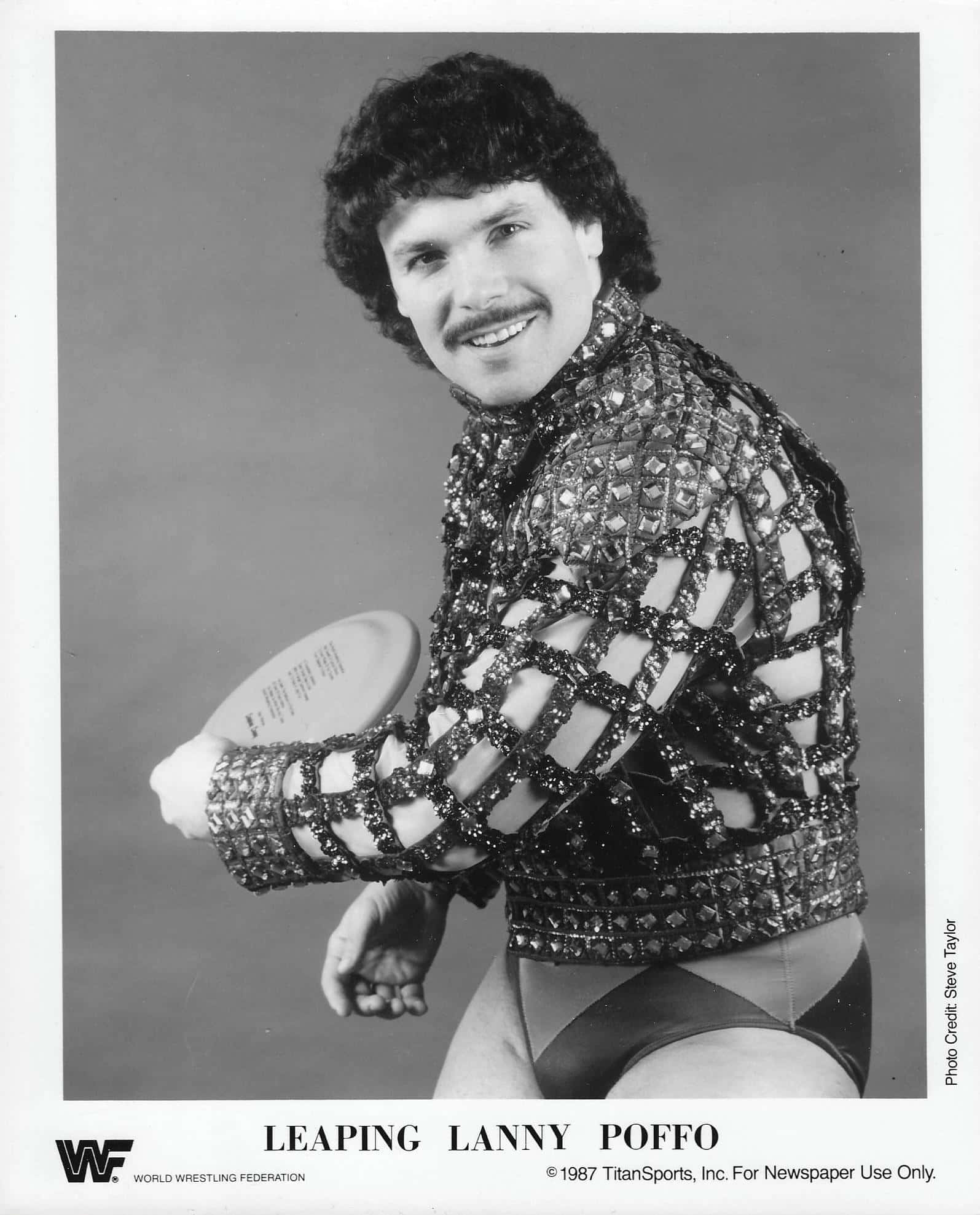摔角 桌布 壁紙 — 摔角手 Lanny Poffo 穿著閃亮網狀服裝，在簡單的攝影棚背景前拿著飛盤的黑白肖像。
