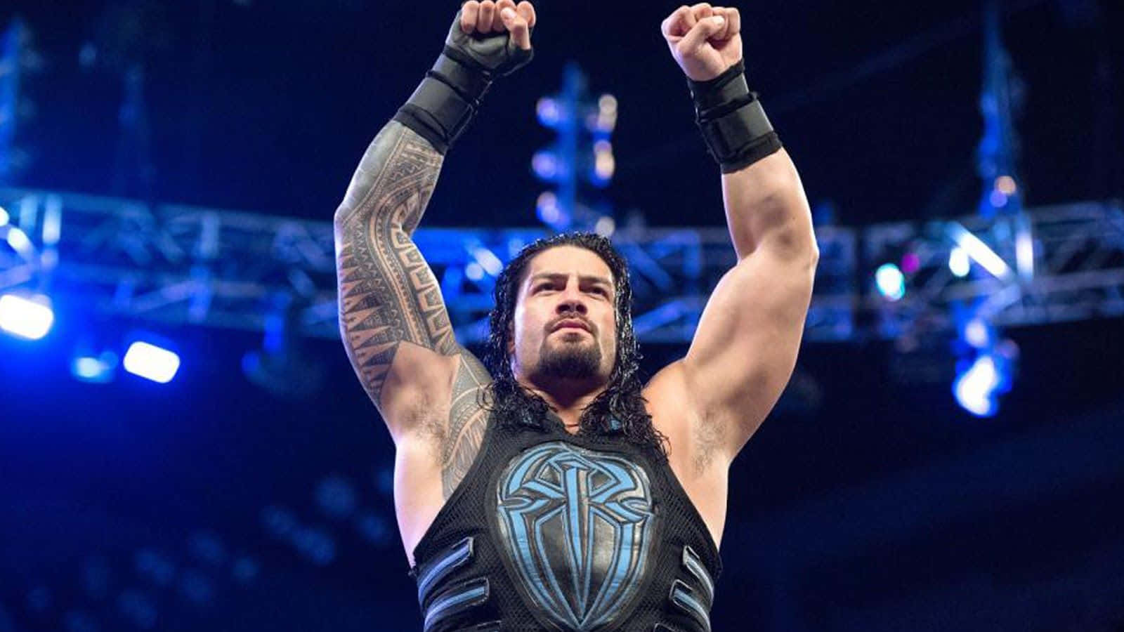 羅曼·雷恩斯 桌布 壁紙 — 摔角手 Roman Reigns 在明亮的藍色場館燈光下高舉拳頭慶祝勝利，背景是模糊的人群。
