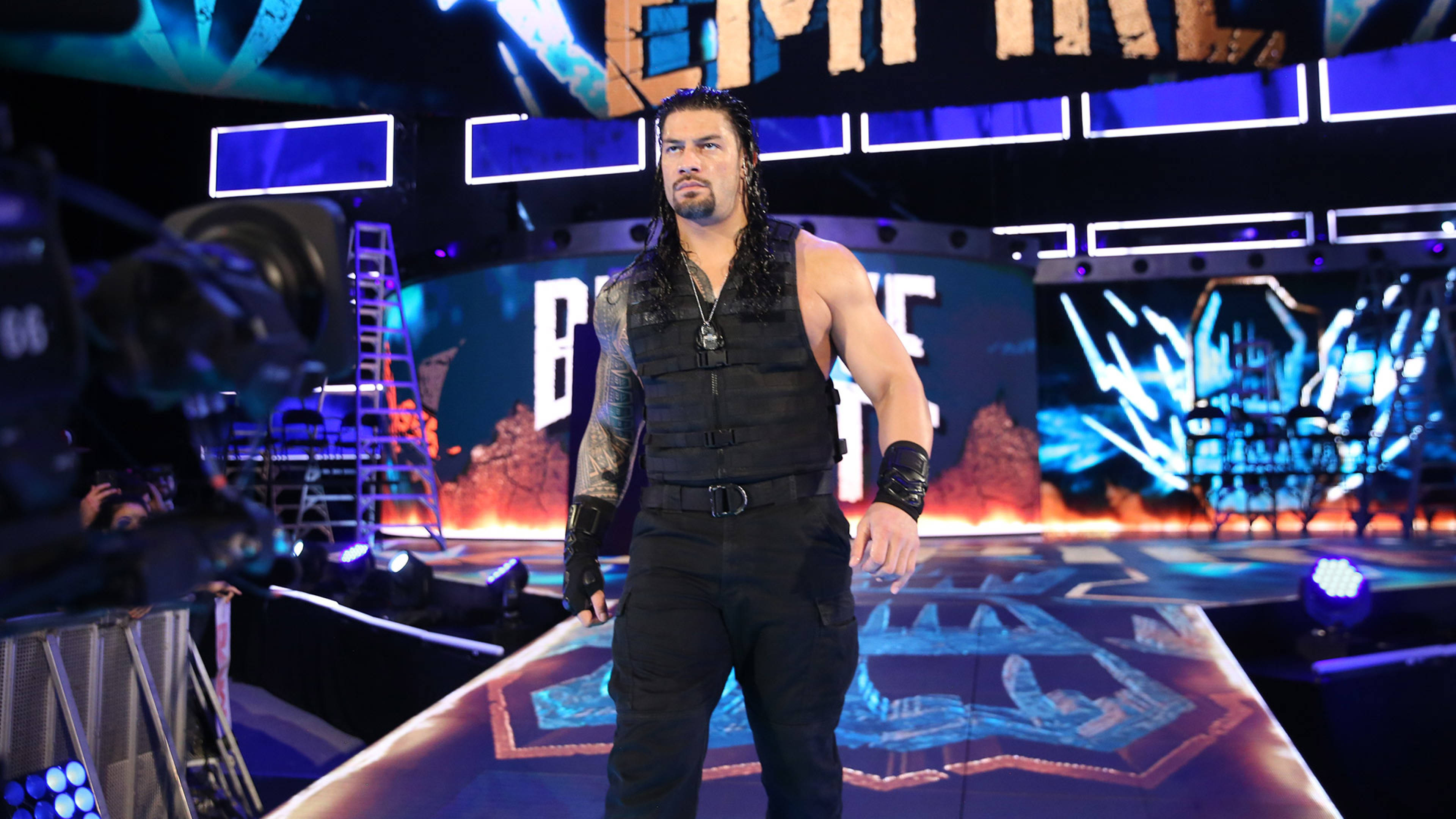 Roman 桌布 壁紙 — 摔角手 Roman Reigns 自信地穿著黑色背心和褲子，站在一個燈光明亮、帶有藍色和橙色燈光的摔角舞台上。