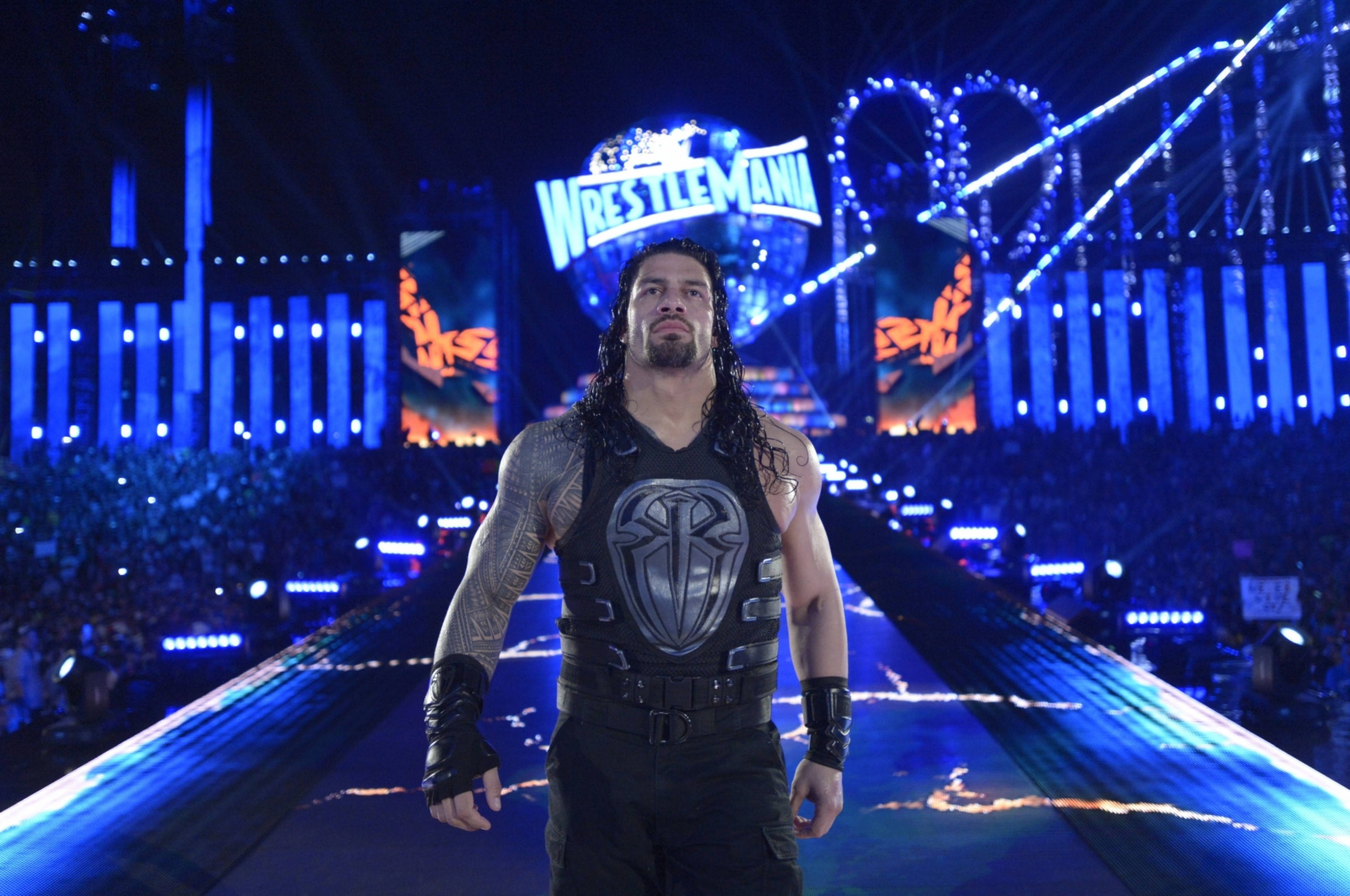 Roman 桌布 壁紙 — 摔角手 Roman Reigns 自信地走在藍色燈光的競技場斜坡上，在明亮的舞台燈光下，身後有 'WrestleMania' 標誌。