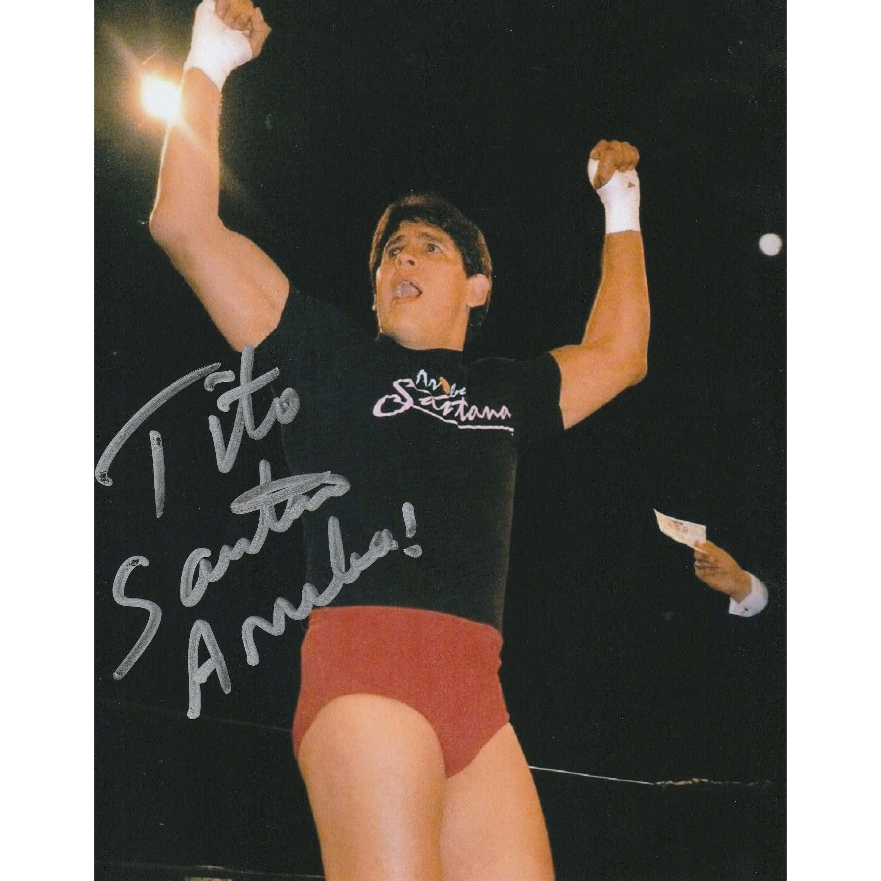 摔角 桌布 壁紙 — 摔角手 Tito Santana 在明亮的舞台燈光下，身穿黑色襯衫和紅色短褲，勝利地高舉雙拳。