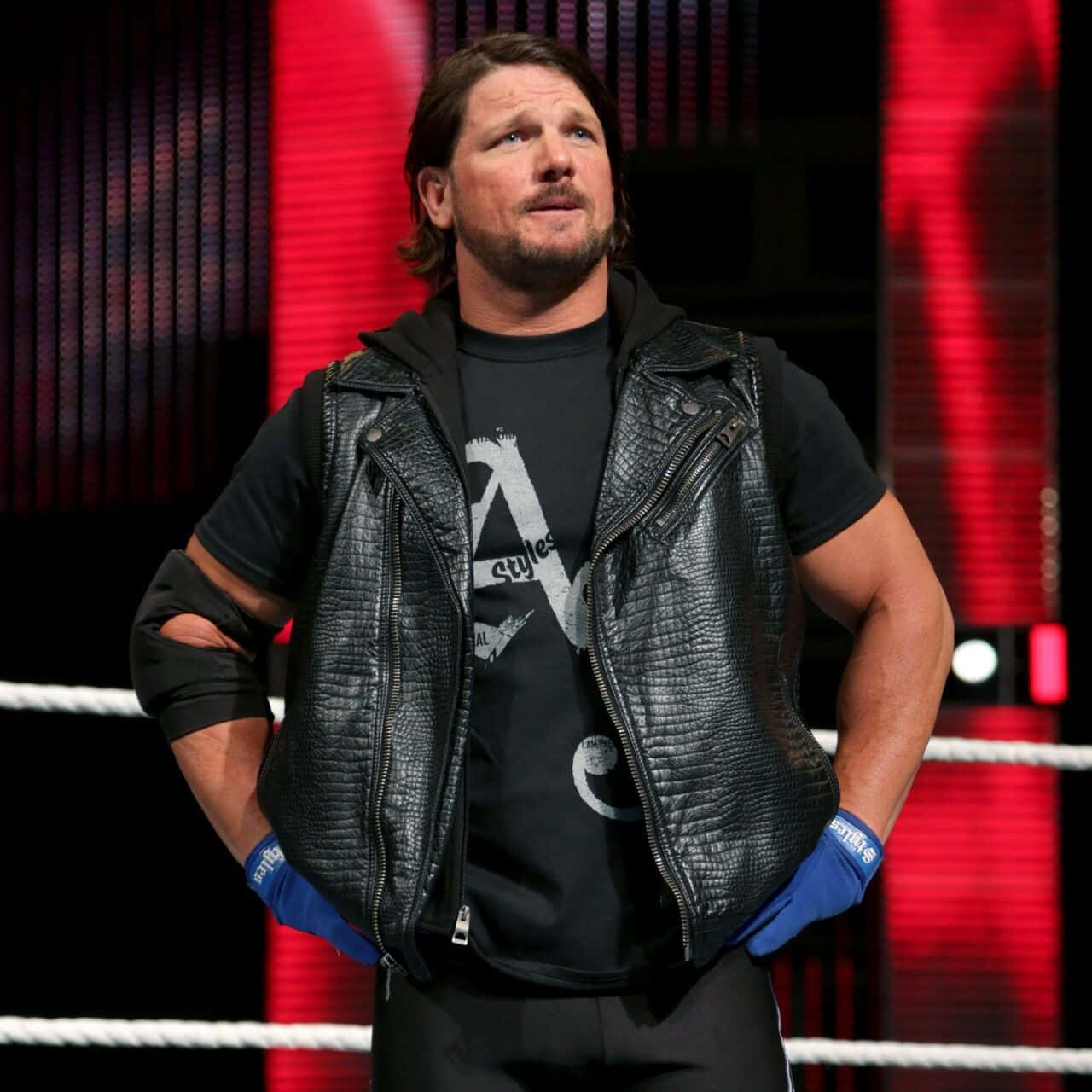 WWE 桌布 壁紙 — 摔角手AJ Styles自信地站在摔角擂台上，身穿黑色皮背心和藍色手套，背景為紅色燈光。